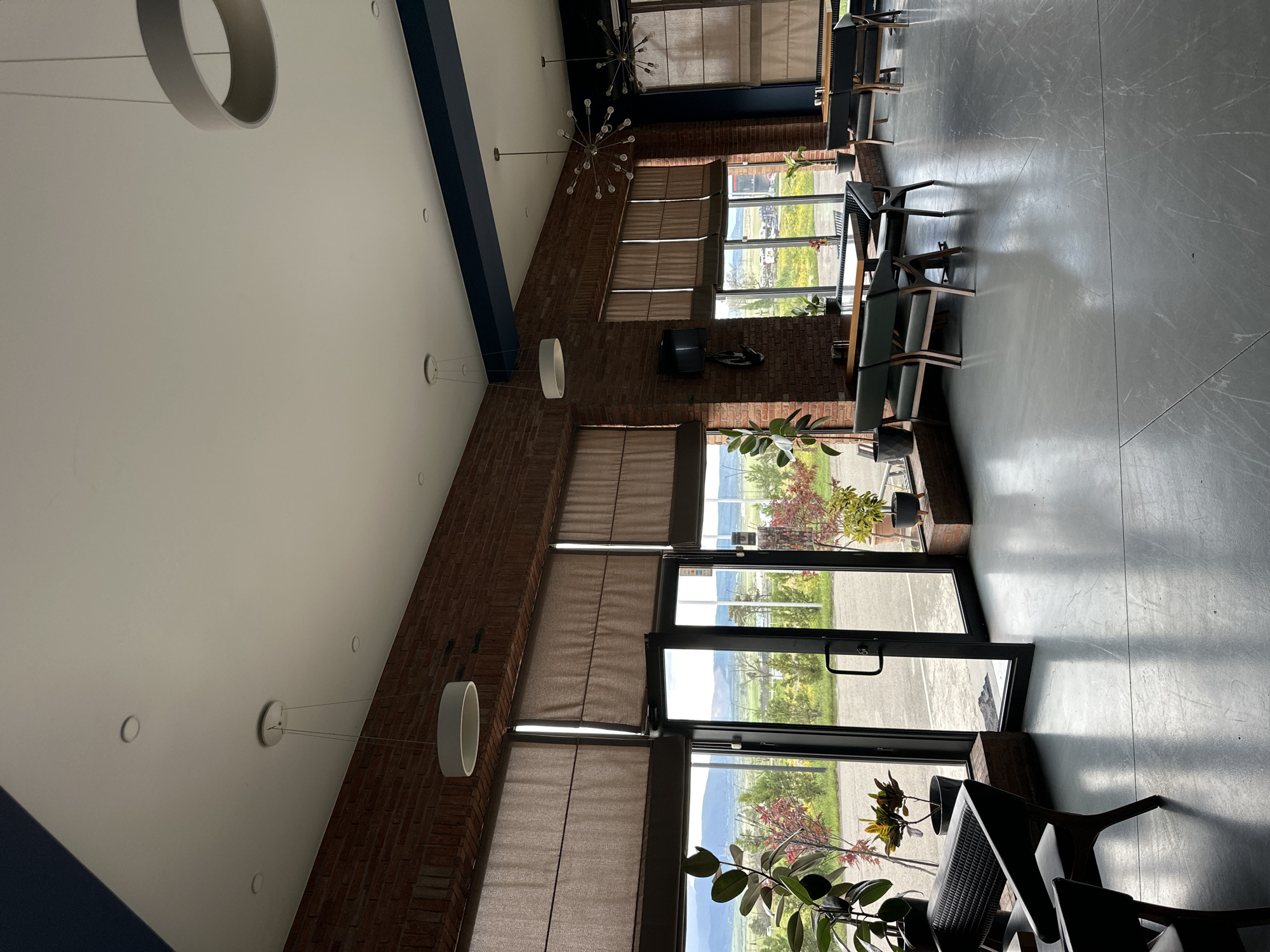 The image captures the interior of a commercial space, possibly a restaurant or cafe, in Marneuli, Georgia. The room features exposed brick walls on the right side, large windows with partially drawn blinds, and a gray concrete floor. Daylight floods the space through the windows, revealing a view of the surrounding landscape. Furniture includes tables and chairs, and potted plants are positioned near the windows. The ceiling is white with modern, minimalist lighting fixtures. The overall impression is of a modern, industrial-chic design aesthetic. There is no evidence of people in the scene.