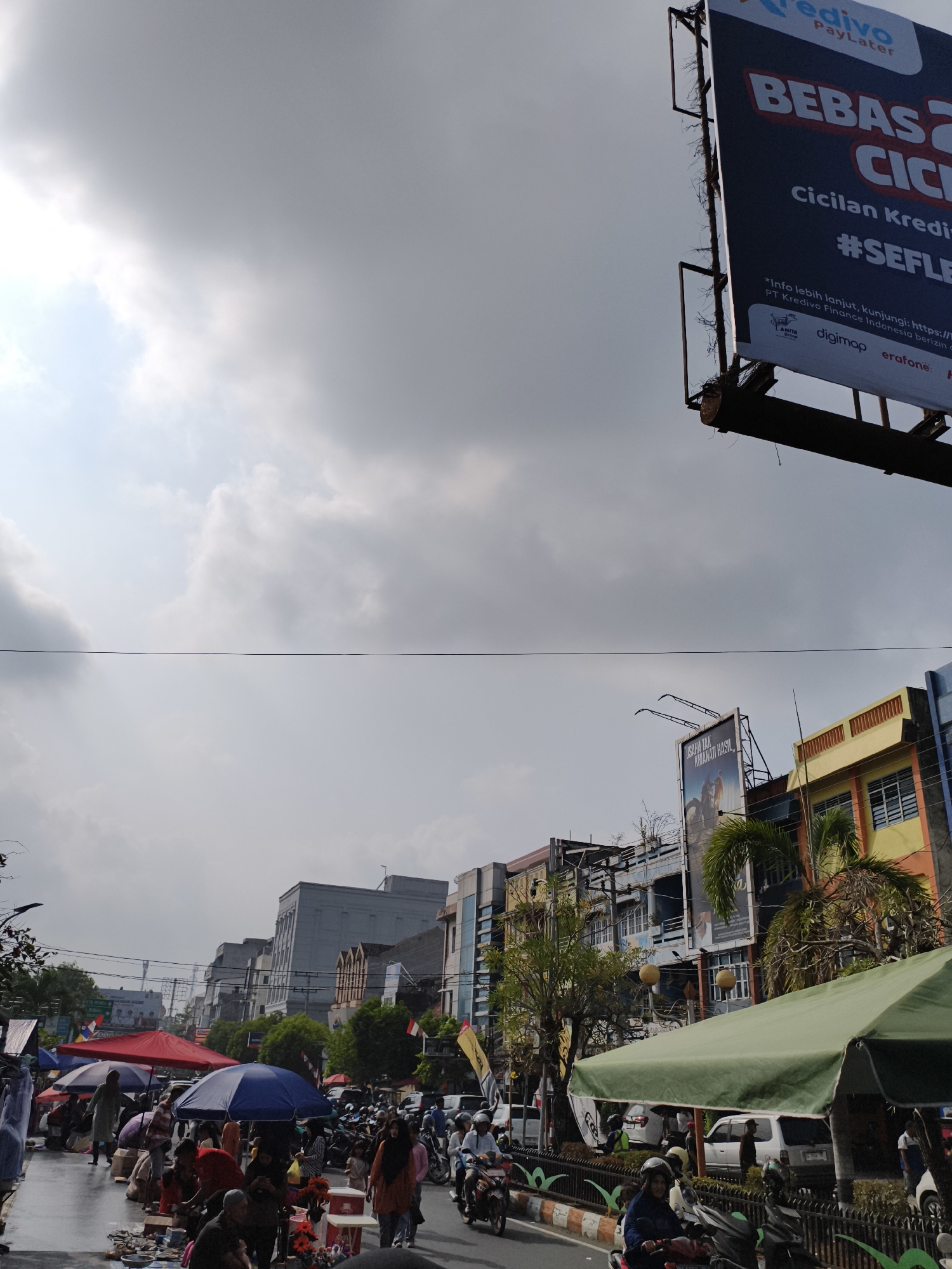 The image captures a bustling street scene in Baturaja, Indonesia, likely during daytime given the bright sky and shadows. In the foreground, a market is in full swing with numerous people browsing and interacting with vendors. Various colorful umbrellas, predominantly red and blue, provide shade for the stalls and shoppers. Items are laid out on the ground for sale.
The street is lined with buildings of varying architectural styles and colors, including a prominent yellow and orange building on the right. Some buildings appear older and somewhat weathered, while others look more modern. A large billboard advertising "BEBAS 2 CICILAN" and "#SEFLE" is visible in the upper right portion of the image, suggesting a commercial area. Another billboard featuring a motorcycle advertisement is also present.
The road itself is paved and shows signs of traffic, with several motorcycles and cars present. People are seen riding motorcycles, indicating local transportation. Power lines crisscross the sky, a common sight in urban Indonesian settings. The overall atmosphere is one of lively commerce and daily activity within the city. The sky is partly cloudy, with a mix of sunlight and gray clouds.