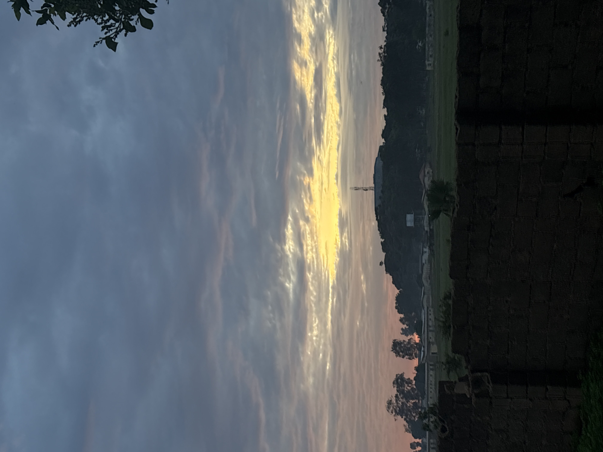 This image depicts an outdoor landscape captured during either sunrise or sunset, characterized by a heavily clouded sky exhibiting gradients of dark blue-grey in the upper portion, transitioning to lighter grey and then distinct streaks of orange and bright yellow at the horizon. The scene is set in Bukuru, Nigeria.
In the background, a prominent, dark green, tree-covered hill or small mountain range is visible, appearing largely in silhouette. Situated atop this elevation are a tall, slender communication tower or antenna and a building structure with a flat roof. To the right of the tower, a white water tank on a raised stand is discernible.
Below the hill, in the midground, several low-rise buildings with light-colored walls and darker roofs are present, interspersed with trees. In front of these structures lies an expansive green field, indicative of agricultural land or open pasture.
The immediate foreground is dominated by a dark, weathered wall constructed from brick or concrete blocks. The wall runs horizontally across the lower part of the frame, displaying an irregular top edge with visible damage and missing sections, particularly towards the left. Minimal sparse green vegetation, consistent with grass or small bushes, is present in the narrow strip between the camera's perspective and the wall, as well as peeking above the wall's uneven top edge. A leafy branch from an unseen tree partially frames the upper right corner of the sky. No human figures or discernible activities are observed within the frame.