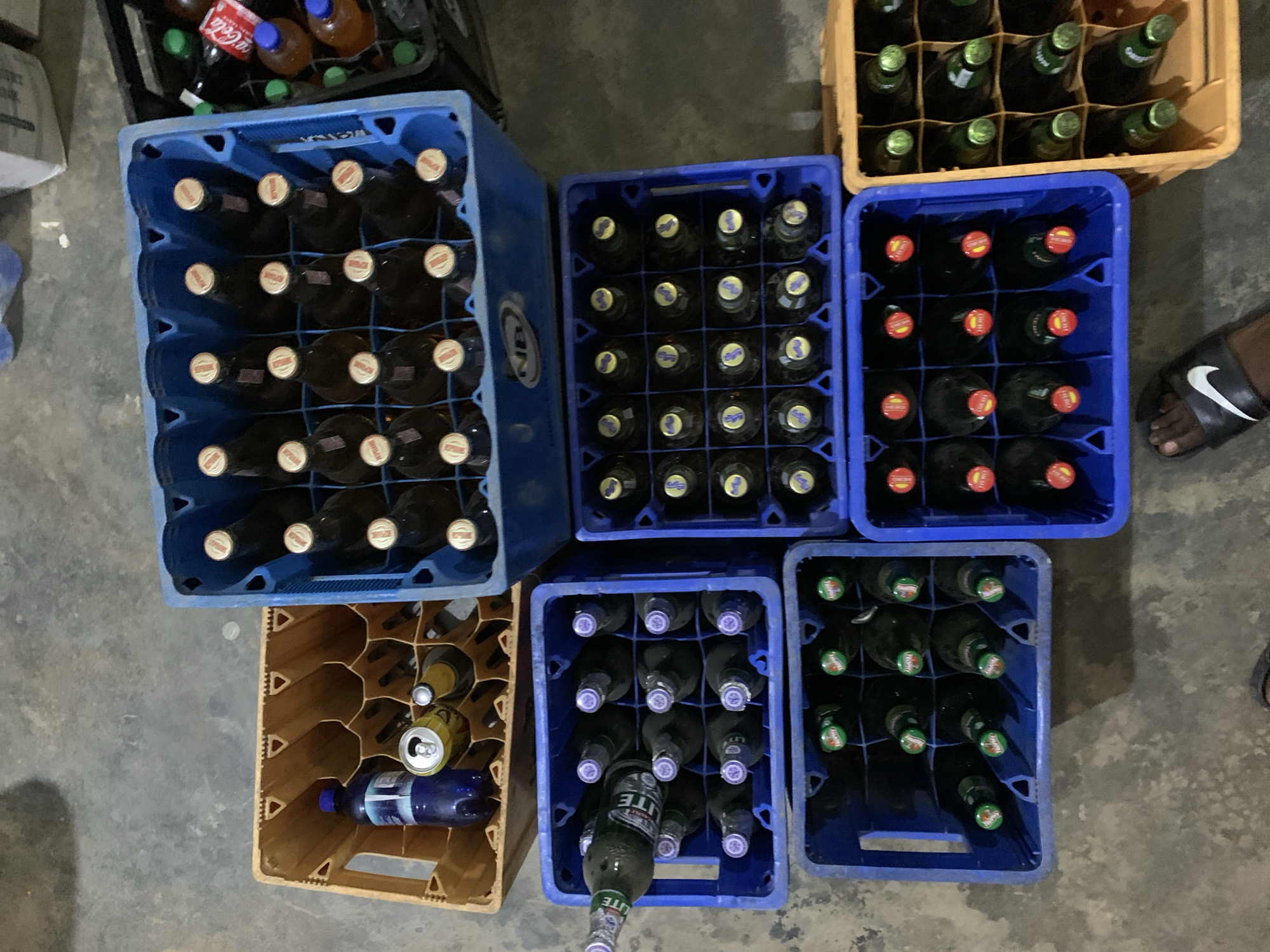This image, captured in New Stadium, Nigeria, depicts an overhead view of eight beverage crates arranged on a grey, concrete floor.
In the upper central area, a large blue plastic crate contains 24 brown glass bottles with yellow and orange crown caps; an "NB" logo is visible on the crate's side. To its immediate left, a yellow plastic crate holds one blue plastic bottle, one open yellow soda can, one brown glass bottle with a white cap, and appears partially empty. Adjacent to the top-right blue crate, a black plastic crate is partially visible, containing various bottles including a red Coca-Cola bottle and several clear plastic bottles with orange liquid.
Below the yellow plastic crate on the left, a blue plastic crate contains 20 green glass bottles with purple crown caps; one bottle is positioned horizontally, and some labels display "STAR LITE." To its right, another blue plastic crate holds 20 dark glass bottles with blue and yellow crown caps.
In the bottom row, three blue plastic crates and one yellow plastic crate are visible. The bottom-left blue crate contains 12 dark glass bottles with green crown caps, some of which display "TROPHY" branding. The adjacent blue crate to its right holds 12 dark glass bottles with red and yellow crown caps, some bearing "HERO" branding. To the far right, a yellow plastic crate contains 12 green glass bottles with green crown caps.
A dark-skinned foot, wearing a black Nike slide with a white swoosh, is visible in the lower-central part of the image. The floor throughout the scene is uniformly grey and appears textured.