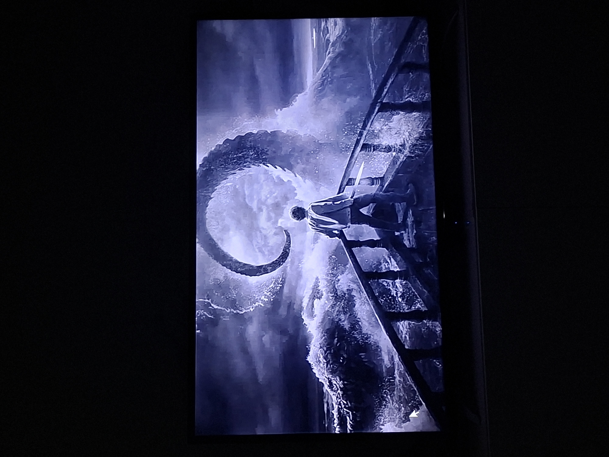 The image depicts a scene displayed on a screen, likely a television or monitor, in a dark room. The scene itself appears to be digitally rendered and portrays a dramatic, high-seas adventure. A man clings to a ladder attached to a ship or structure, amidst a turbulent ocean. A giant, swirling tentacle emerges from the water, seemingly threatening the man. The overall tone is dark and ominous, with a monochrome color palette dominated by shades of blue and grey. The scene appears to be a frame from a movie or video game. There is no indication of Galabovo, Bulgaria.