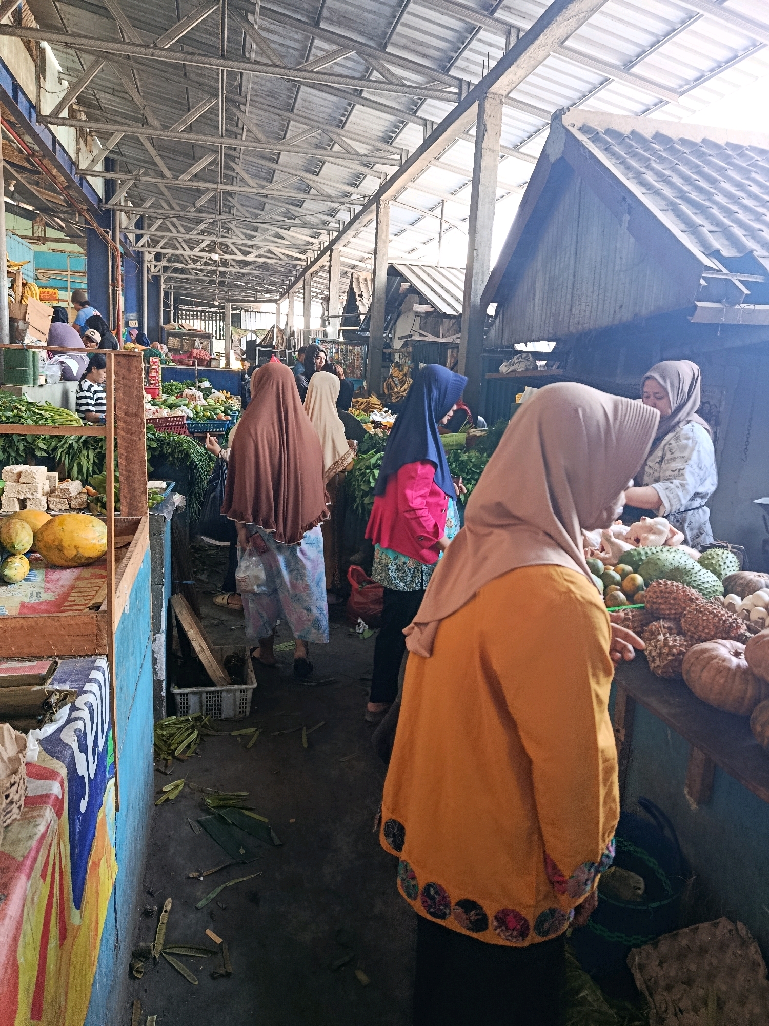 In Bululawang, Indonesia, a bustling market scene unfolds under a metal-framed, corrugated roof structure. The open-air market features stalls laden with fresh produce. In the foreground, a woman wearing an orange headscarf and tunic stands with her back to the camera, facing a stall filled with various fruits and vegetables, including what appear to be pumpkins, durians, and citrus fruits. To her left, a stall displays papayas and other fruits. Further into the market, several other women, mostly wearing hijabs, are visible shopping and interacting with vendors. One woman, in a white shirt and gray headscarf, appears to be a vendor, attending to customers at a stall piled with chickens and produce. The ground is a mix of dirt and scattered leaves and plant matter. The overall impression is one of lively commerce in a local Indonesian market.