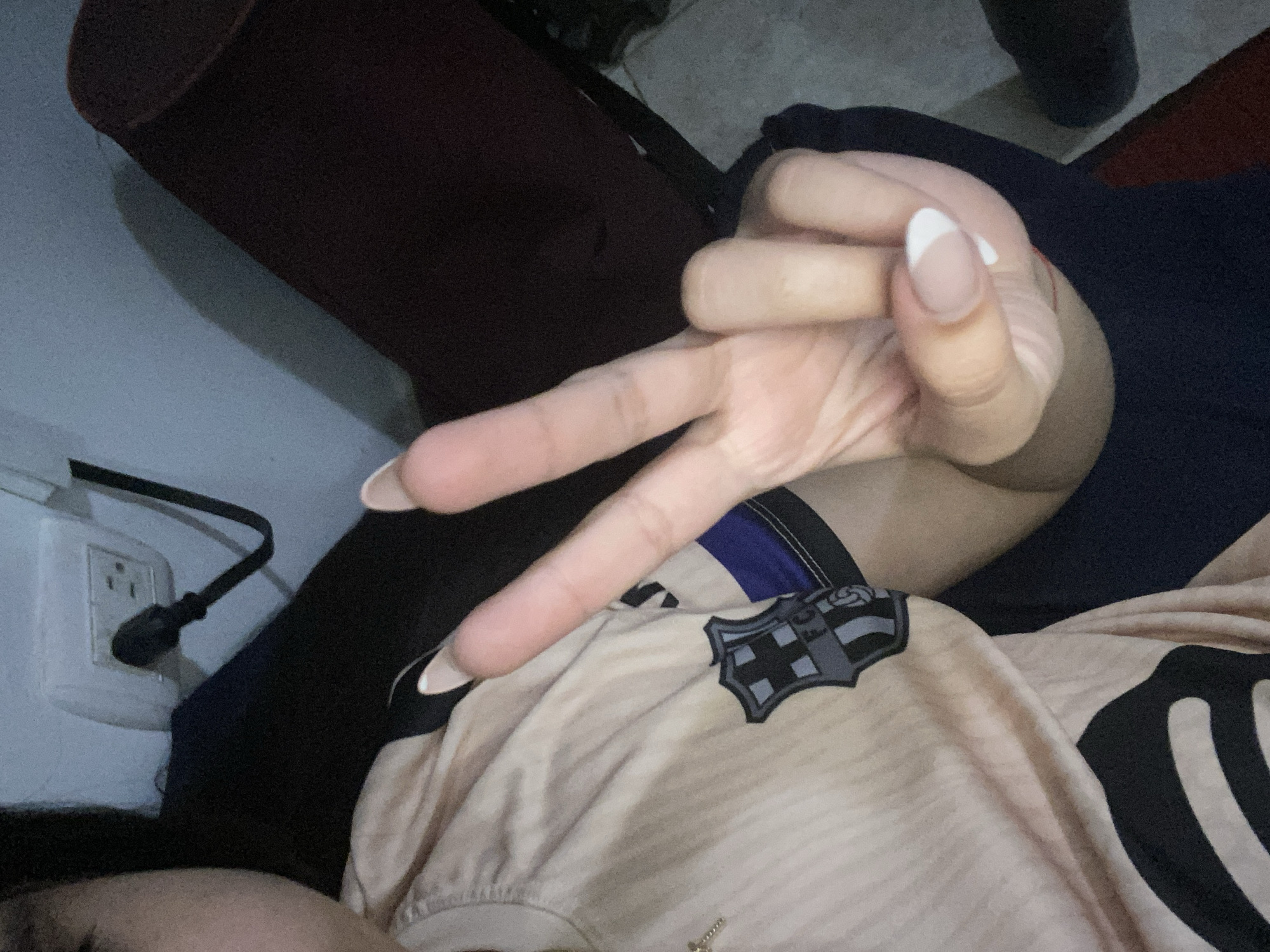 The image shows a person wearing a light beige shirt with the Barcelona FC emblem, making a peace sign with their hand. The hand has a French manicure. The person is sitting on what appears to be a dark blue or black soft surface with a dark red or maroon object behind them. A wall outlet and cord are visible in the background. The location is not identifiable as being in Cotoperíz, Venezuela, or any other specific location based on the visible elements.