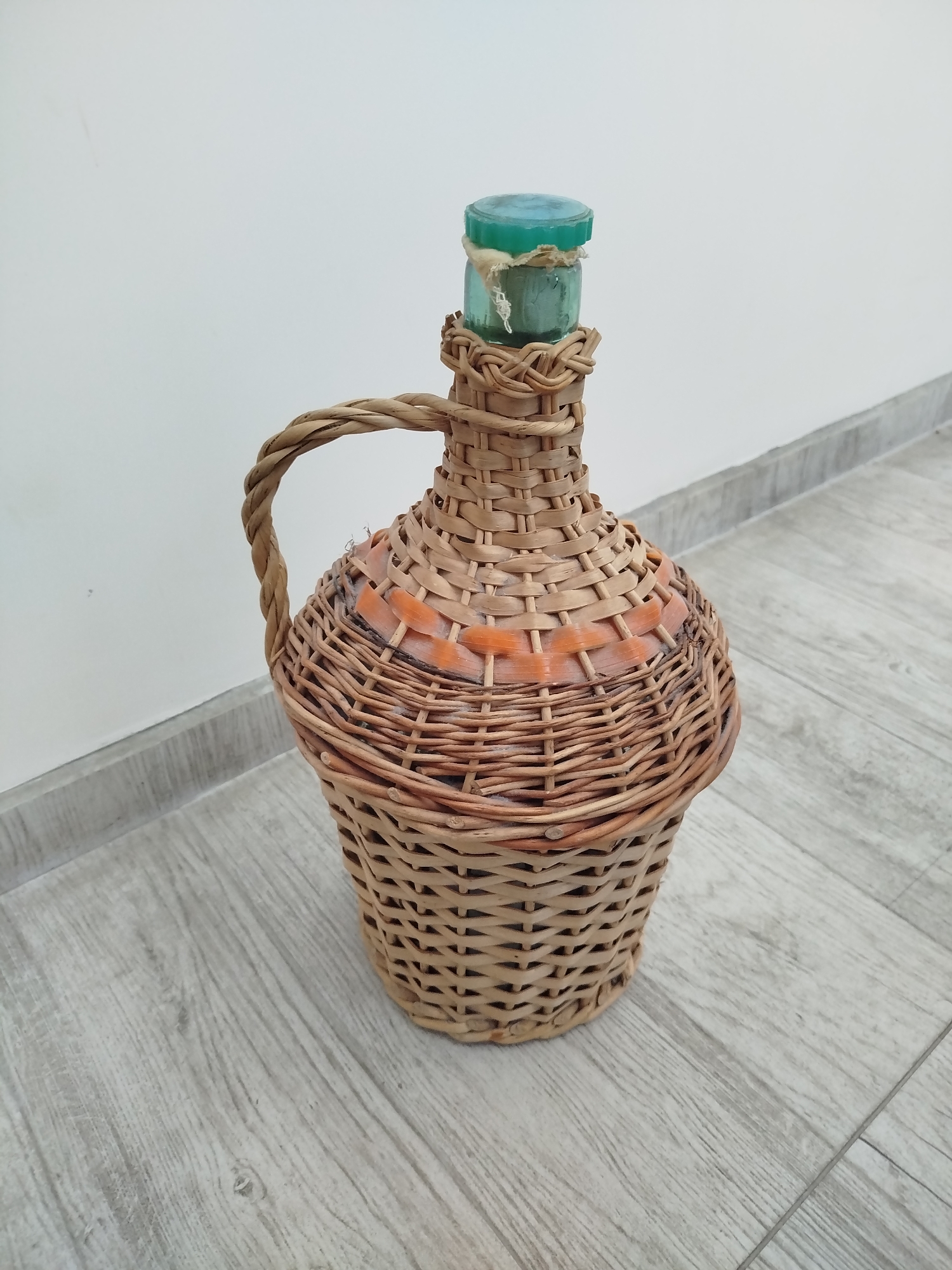 The image shows a wicker-covered glass bottle, likely a demijohn, sitting on a light-colored wood-grain floor. The bottle has a green glass top. The wicker casing is woven around the bottle, forming a handle and base. There is a white wall in the background with a grey trim. The location is Varna, Bulgaria.