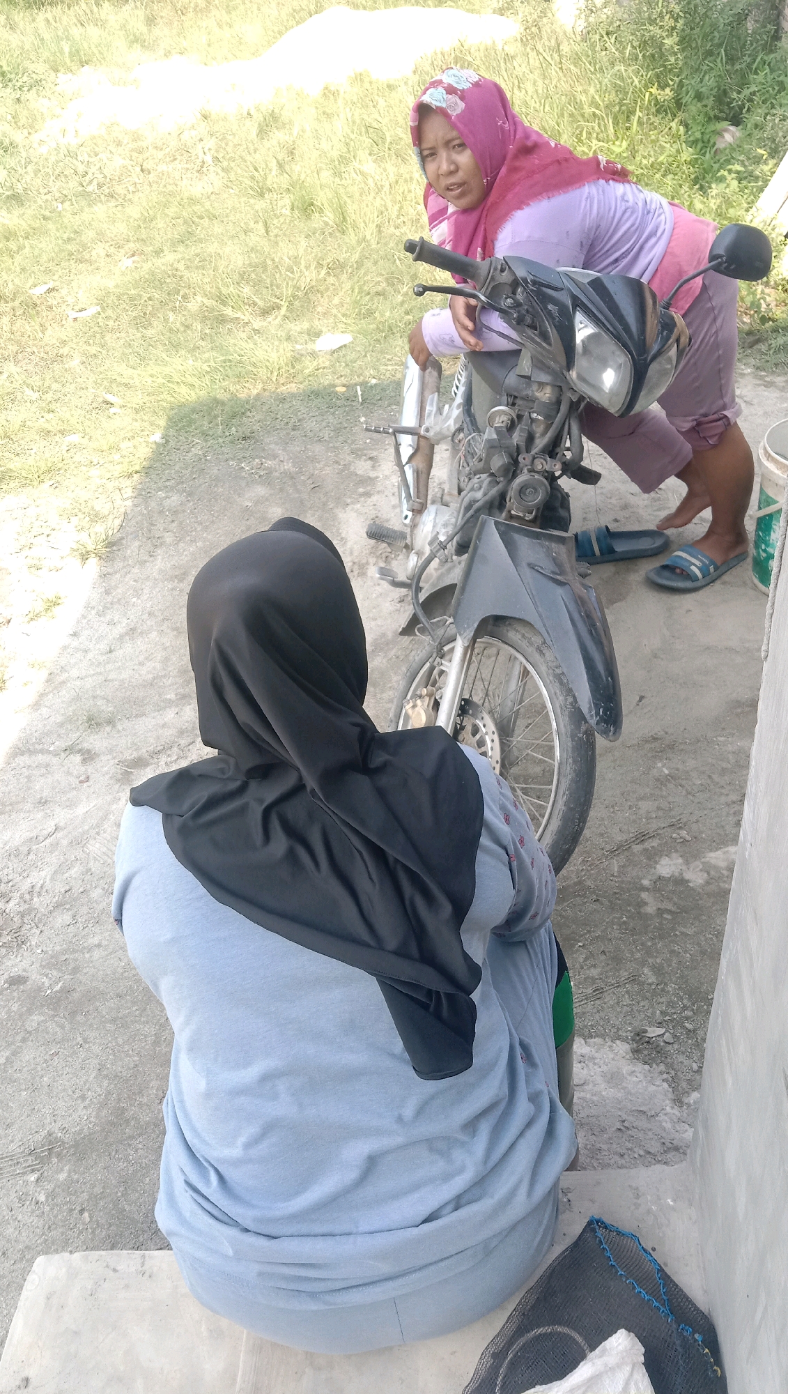 Two women, one wearing a pink hijab and a purple top, and the other wearing a black hijab and a light blue top, are near a motorcycle. The woman in the pink hijab is leaning over the motorcycle and looking towards the camera. The woman in the black hijab is facing away from the camera, with her back to the viewer, sitting on a step. The scene appears to be outdoors, possibly in a rural area or a less developed part of Kota Pari, Indonesia, with dry grass and a dirt or concrete ground. The lighting suggests it is daytime.