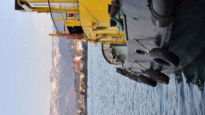 The image depicts a harbor scene in Batumi, Georgia. The foreground is dominated by the starboard side of a partially visible vessel, likely a tugboat, positioned at an angle with its bow facing left and into the water. The boat's hull is dark grey, equipped with a series of black rubber tires serving as fenders, secured by ropes and chains. Its upper structure is painted bright yellow, featuring a cabin with visible windows, a doorway, and a metal railing along the deck. Nautical equipment, including dark bollards wrapped with thick green and brown ropes, and red lifebuoys, is present on the yellow deck section.
The midground consists of choppy, dark blue-green water. Two small, reddish-orange buoys or boats are visible on the water further into the distance.
The background reveals a cityscape or port area on the far side of the water. Prominent features include multiple red-orange gantry cranes, indicative of industrial port operations, and a large white vessel docked or operating within the port. Beyond the urban and industrial elements, a range of mountains with visible snow patches on their peaks stretches across the horizon. The sky is a clear, light blue. No individuals are visible, and no dynamic actions or interactions are depicted.