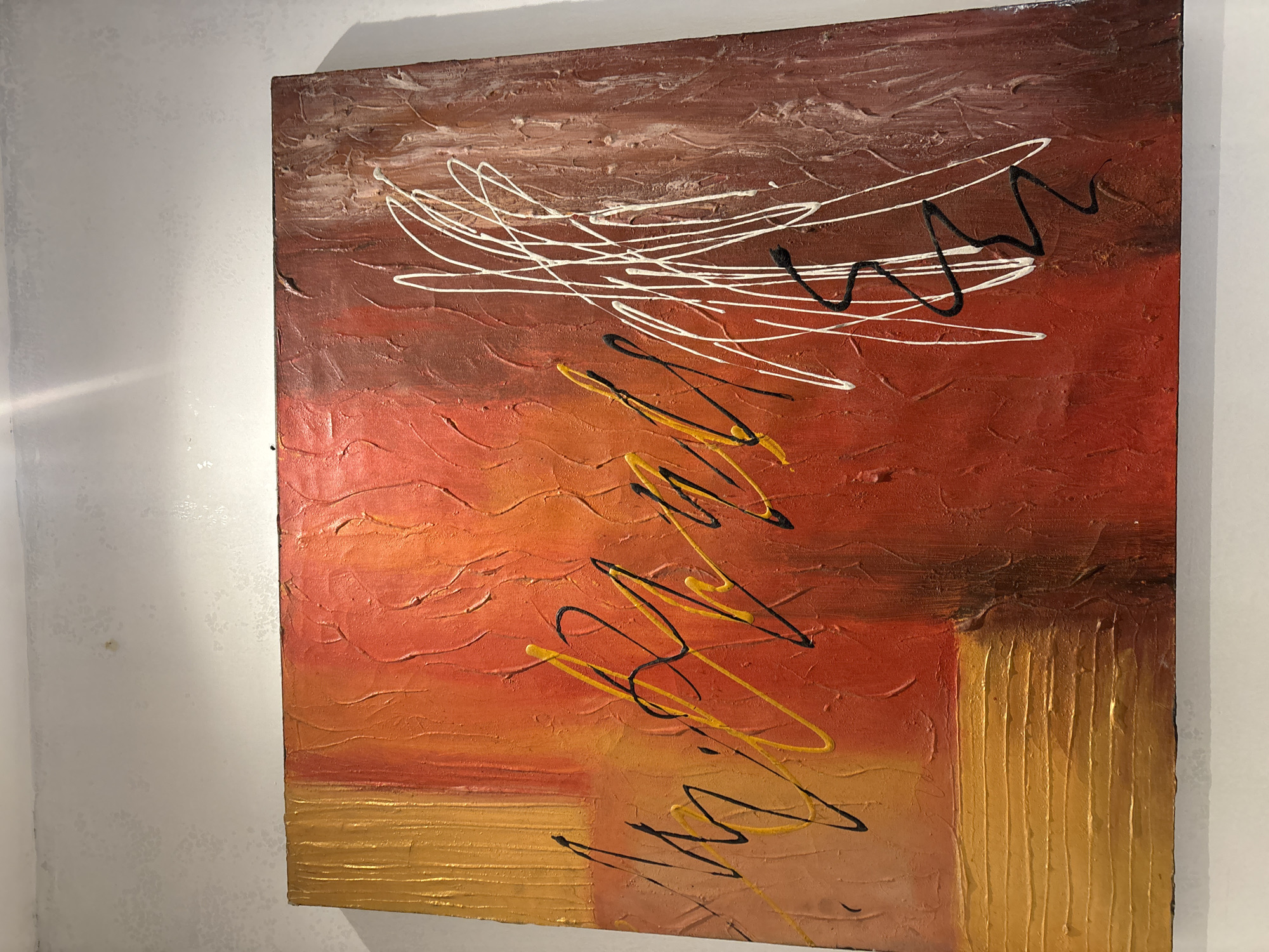 The image shows a textured abstract painting with warm colors like orange, red, and yellow, accented by white, black, and yellow linear elements. The painting is mounted on a wall. The wall has a textured, mottled gray-white finish. The painting is illuminated by a light source from above, creating a bright spot on the upper portion of the wall and casting a slight shadow on the top edge of the artwork. The composition of the painting features bold, gestural strokes and areas of impasto, suggesting a dynamic and energetic aesthetic. The overall impression is one of an indoor setting, possibly a gallery or home, but no specific location in Pekanbaru, Indonesia, can be determined from the visual information provided.
