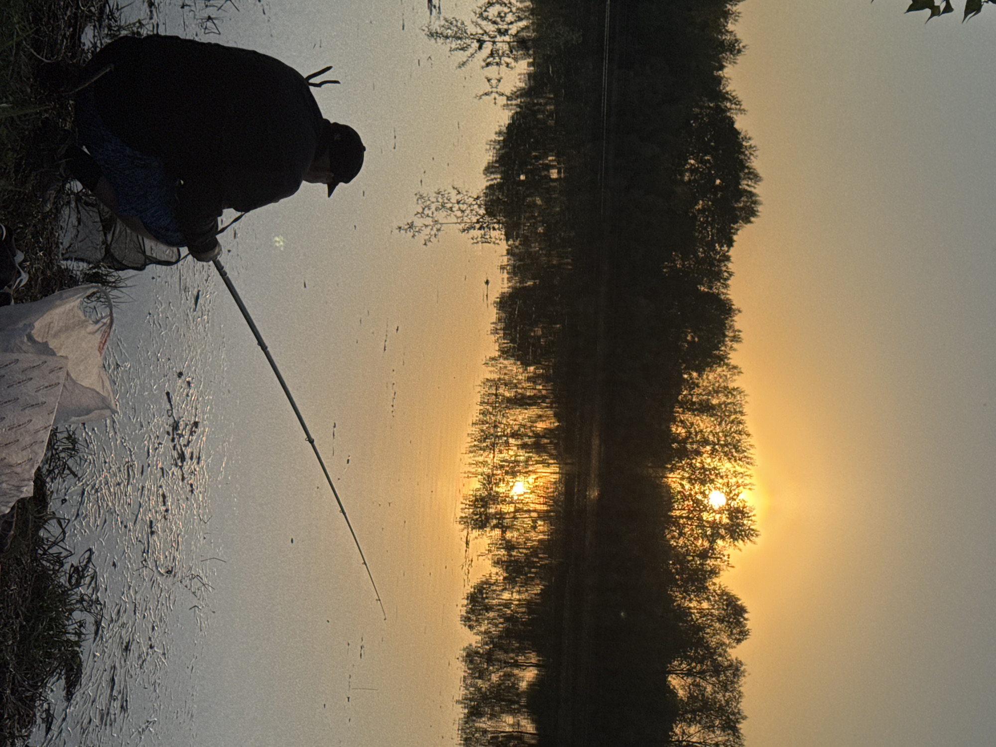 A man is fishing near a body of water that is reflecting the trees and sunset. He is wearing dark clothing and a cap, and he is holding a fishing rod. There is a net and a white bag near him. The location is Chaikovsky, Russia.