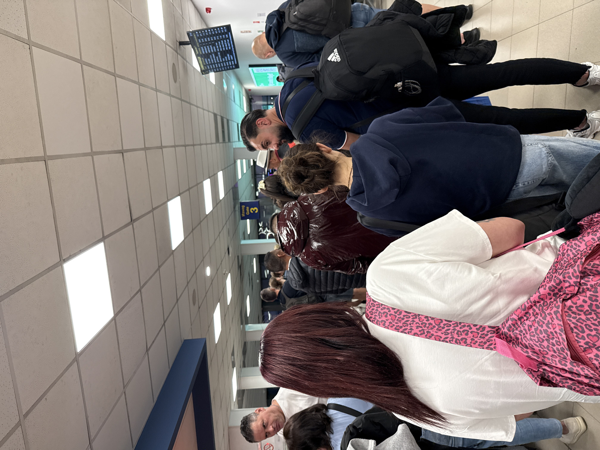 The image depicts a crowded indoor public space, likely an airport terminal in Sofia, Bulgaria, with multiple individuals forming a queue. The perspective is from within the line, looking towards its front.
In the foreground, the back of a person with long, dark red hair is visible, wearing a white top and a distinctive pink leopard-print strap of a bag across their shoulder. To their left, the top of a man's head with grey hair and a light blue shirt is seen, alongside another person with dark hair.
The midground features several individuals. A man with a beard and dark hair, wearing a navy blue polo shirt and a black backpack, is prominent. Next to him, the back of a person wearing a dark blue hoodie is visible. Further along the queue, other people are seen, one wearing a shiny maroon jacket and another a black puffer jacket. Many individuals are carrying backpacks, with one clearly displaying an "adidas" logo.
The setting includes a tiled floor and a tiled ceiling featuring numerous square fluorescent light panels. Along the wall, two black digital screens, likely displaying flight or gate information, are mounted. A red dome-shaped device is affixed to the ceiling. A blue sign, partially visible, reads "GATE 3", reinforcing the airport environment. The primary activity is people standing and waiting in line. The time of day is indeterminate due to the artificial indoor lighting.