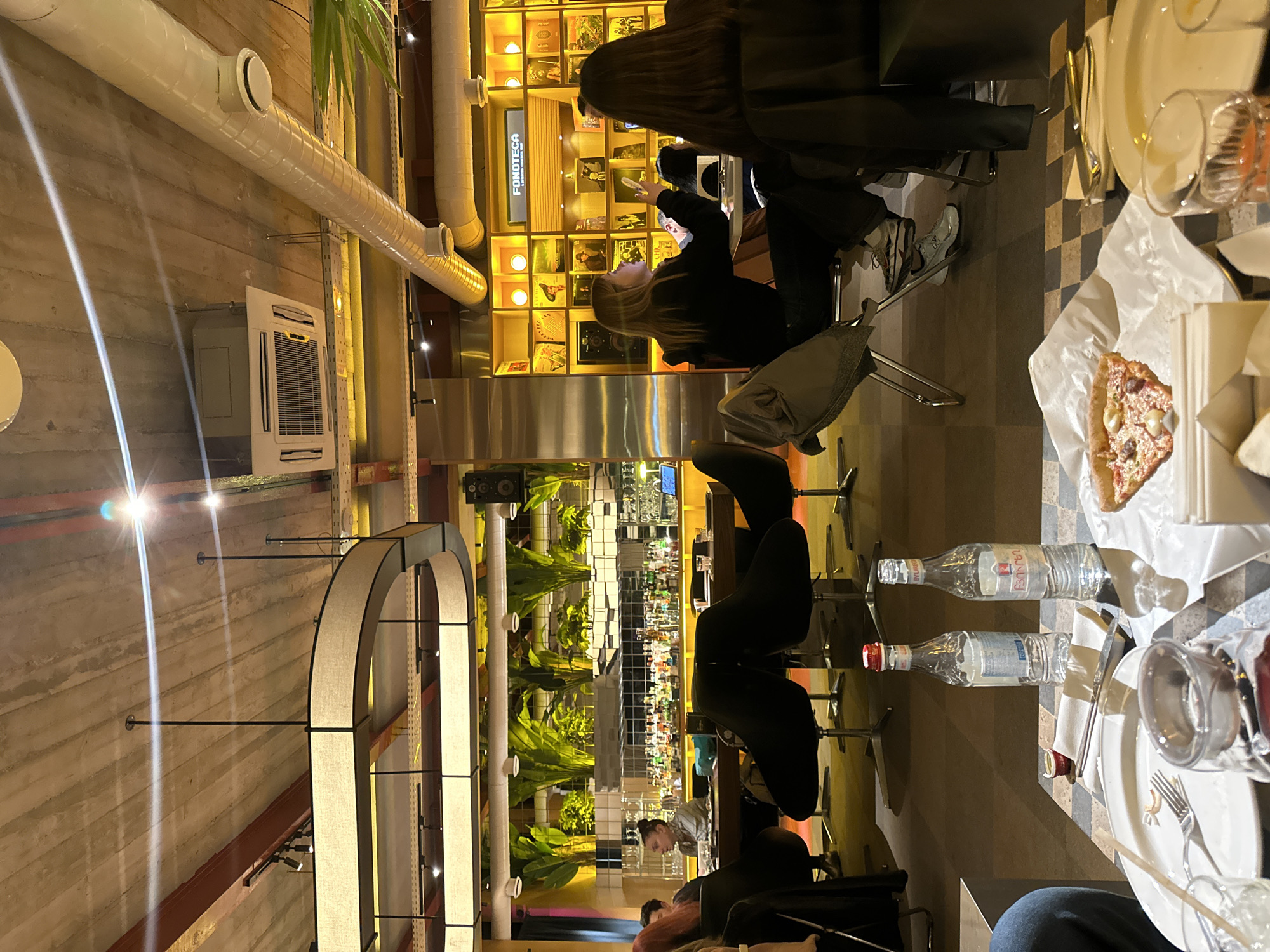 The image depicts the interior of a restaurant in Yerevan, Armenia. The space has an industrial-chic aesthetic, featuring exposed concrete walls, ventilation ducts, and arching light fixtures. A bar area is visible with a mirrored backsplash and plants along the counter. Diners are seated at tables, some with pizza, bottled water, and place settings. People are eating, taking pictures, and interacting. A display case with illuminated compartments is visible in the background.