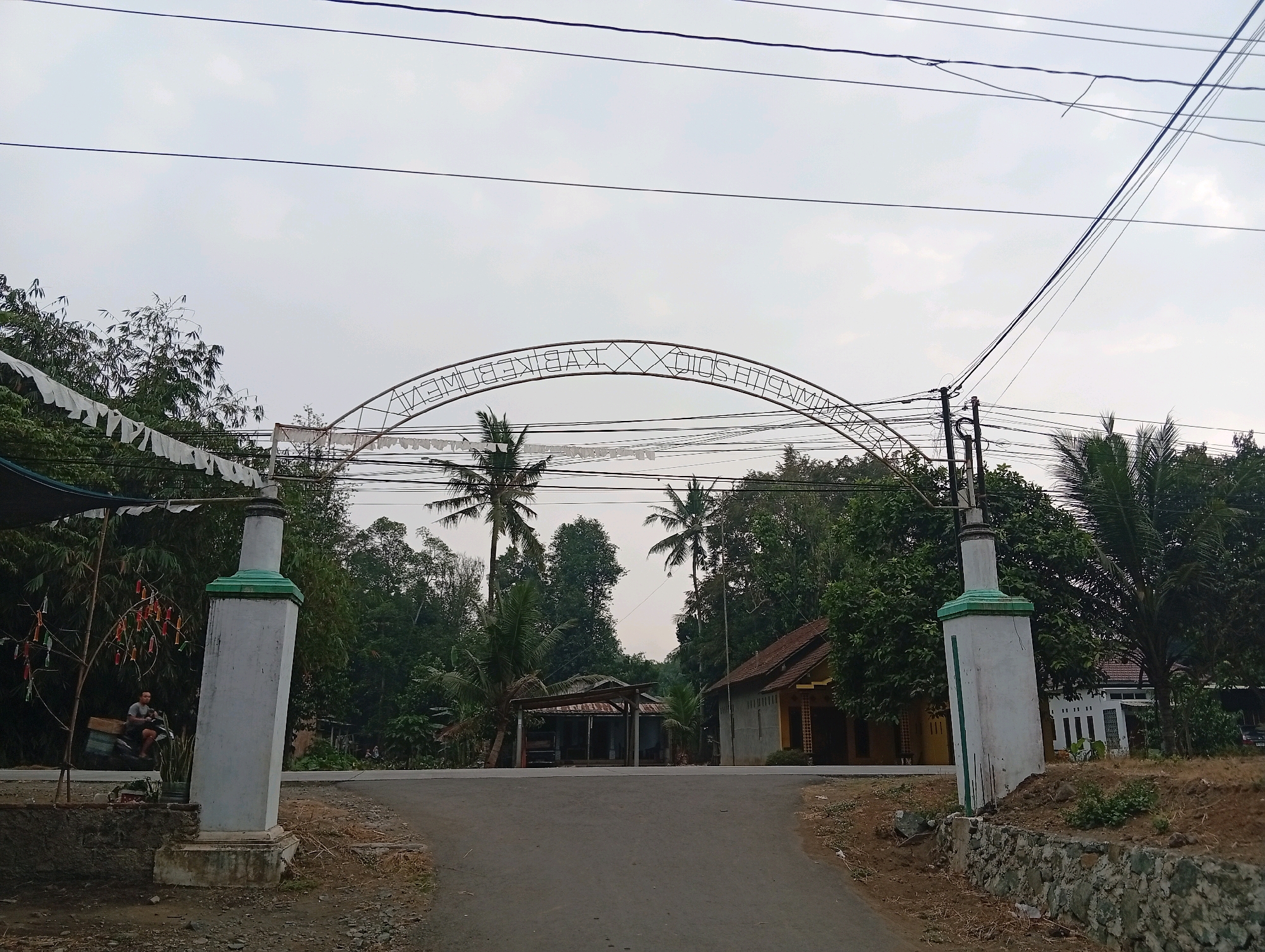 The image shows an outdoor scene on a slightly overcast day in Plempukankembaran, Indonesia. An archway stands prominently in the foreground, with two white pillars topped with green accents. The arch has lettering that appears to be "KWAKEMBAN". White banners or flags hang across the arch. A paved road leads through the archway into a more rural area with lush green trees and vegetation.
To the left of the arch, a person is visible riding a motorcycle with a cargo box. A small tree with colorful decorations hangs nearby. On the right side of the road, a stone retaining wall is visible, and behind it, a small, rustic house with a tiled roof. Further in the background, more houses and trees are scattered, and several power lines stretch across the sky. The overall atmosphere is tranquil and suggests a village or rural entrance.