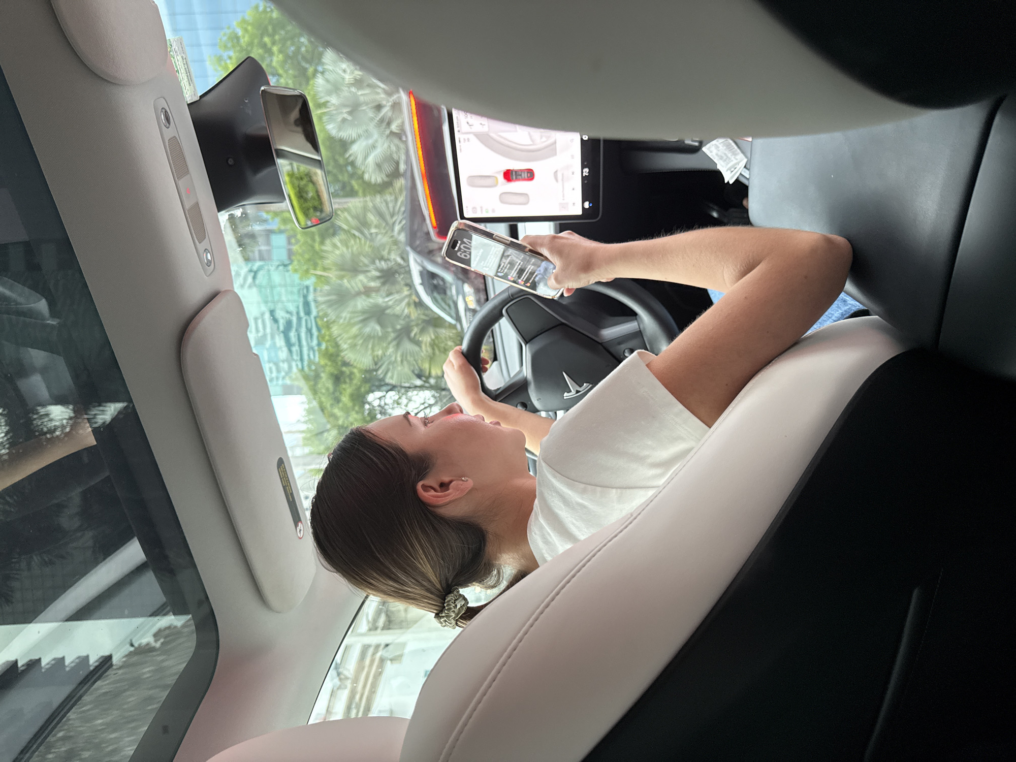 A woman with brown hair pulled into a ponytail is driving a car with a panoramic sunroof. She is wearing a white t-shirt and blue jeans, and is holding a smartphone in her right hand, looking at it. The car has a large touchscreen display in the center console, which shows a car icon and a speed indicator. Outside the car, palm trees and buildings can be seen, suggesting a tropical urban environment. The scene appears to be in Miami, Florida, United States of America, given the distinctive palm trees and modern architecture. The overall lighting and reflections suggest it is daytime.