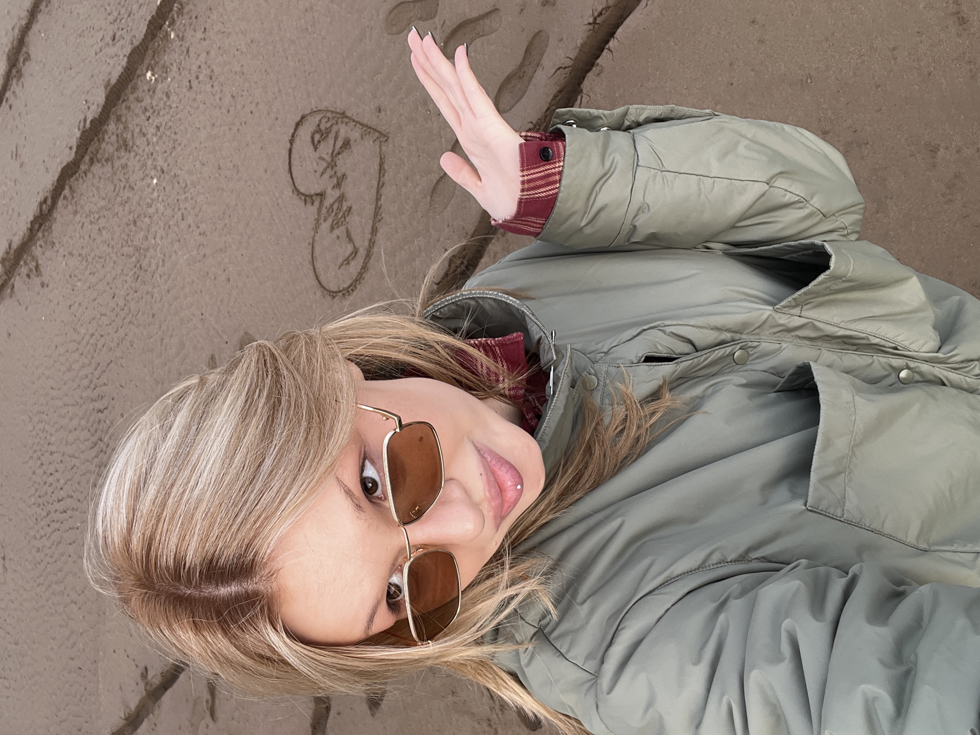 The image shows a woman lying on a sandy surface, possibly a beach. She is wearing sunglasses and an olive green puffer jacket with a red and black plaid shirt visible underneath. Her blond hair is parted and styled. She is holding up her hand, palm facing out. On the sand near her, there is a heart drawn with text written inside. There are also some footprints on the sand. The location is not identifiable as Noviy, Russia, from this image.