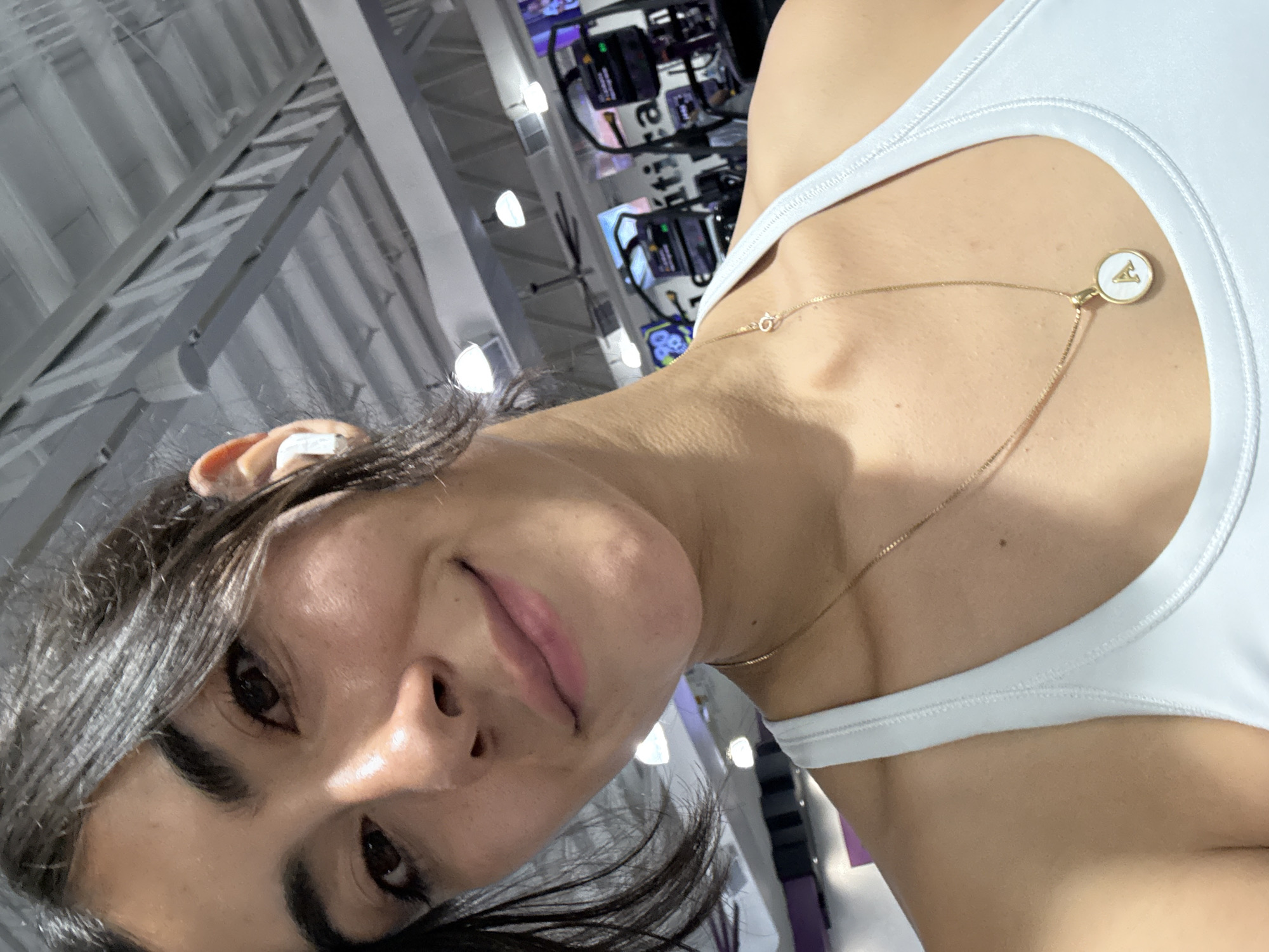 A selfie perspective of a woman with dark hair and brown eyes, wearing a white athletic top and a gold necklace with a round pendant displaying the letter "A". She has an earbud in her left ear. The background reveals a gym setting with various exercise machines. The location is potentially in the city of Puebla, Mexico, inferred from the metadata.