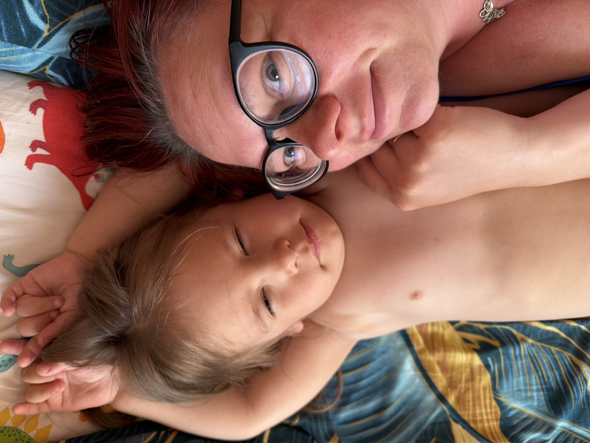 A top-down shot shows an adult with red hair and glasses and a child lying on a bed. The adult's head is at the top of the frame, and the child's head is at the bottom. The adult is wearing black-rimmed glasses. The child has light brown hair and is lying on their back with their eyes closed. The bed has a patterned pillow with a red dinosaur-like figure and a blue and gold palm leaf patterned bedspread. It is not possible to determine if this is in Chaikovsky, Russia, as there are no identifying elements.