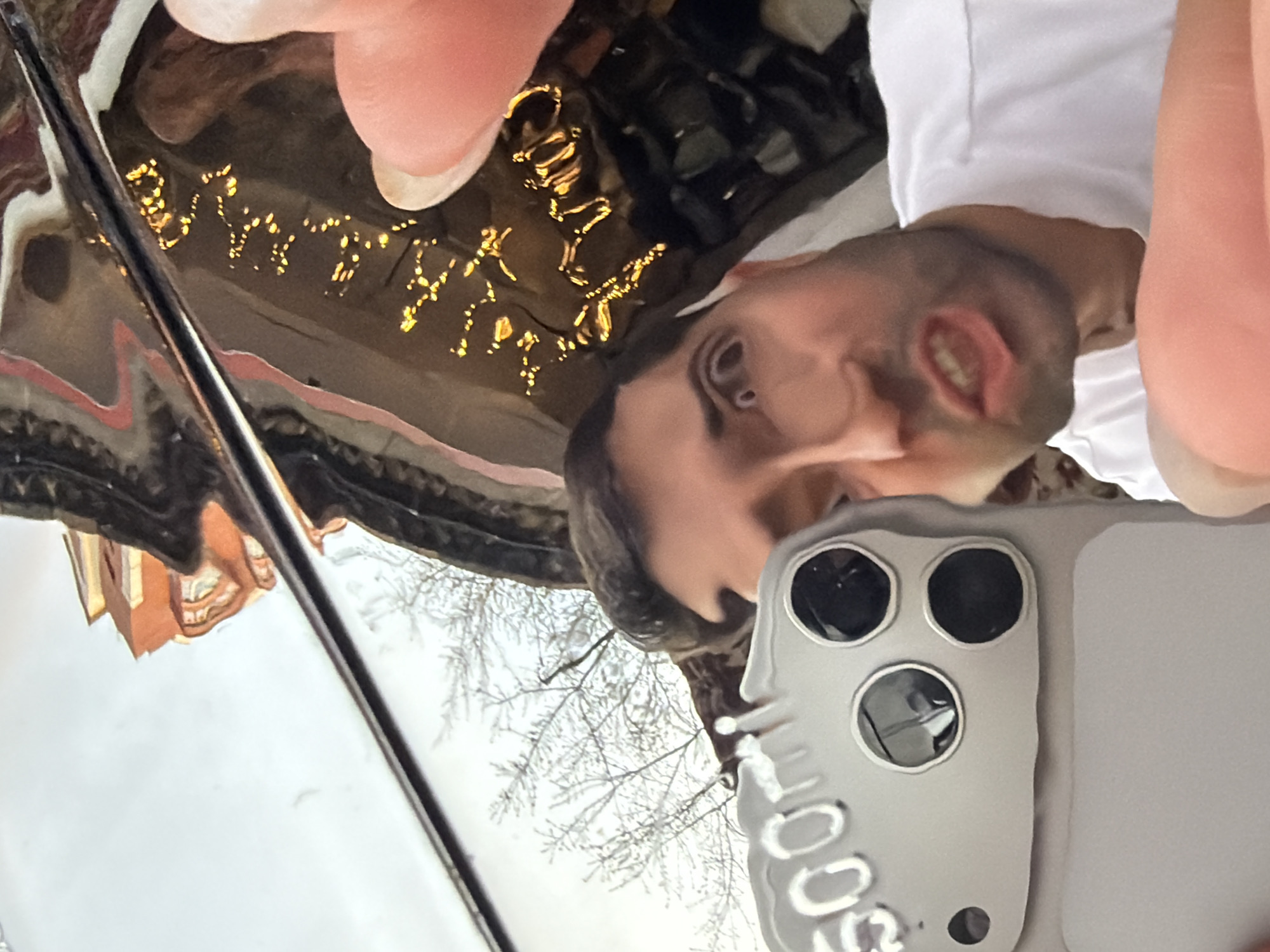 A man is taking a selfie with his phone. The reflection in the car's glossy surface reveals a building and trees, likely in Krasnoyarsk, Russia. The building has a red brick facade with white accents. Bare branches of trees are visible against a pale sky. A finger is visible at the top and right corner of the image. He is wearing a white shirt. There are other items reflected, including some that appear to be gold.