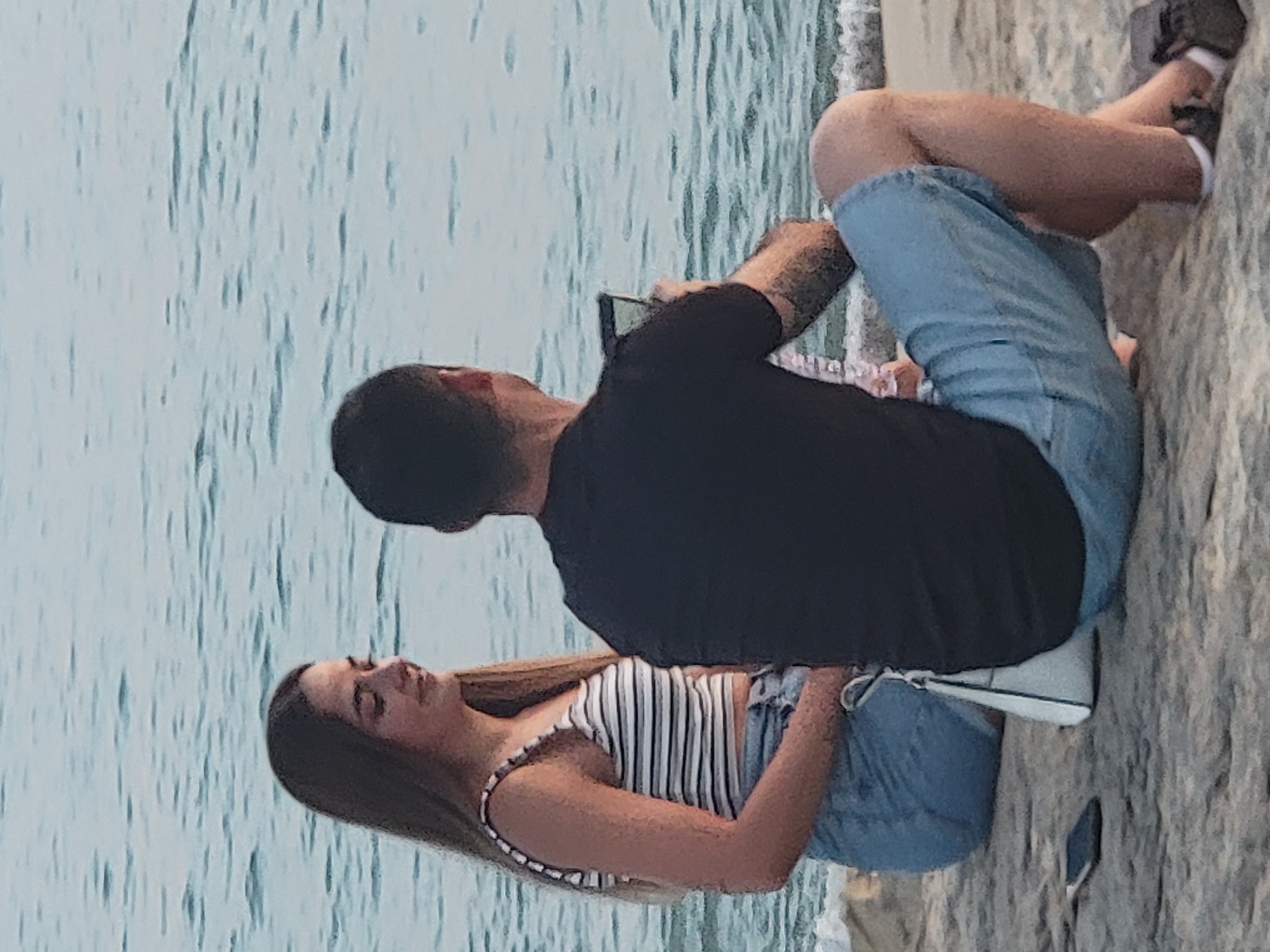 Two individuals, a female and a male, are seated on a rocky surface facing a body of water that appears to be the ocean. The female, on the left, has long brown hair and is wearing a striped crop top and denim shorts. She has a white purse resting on her lap. The male, on the right, is wearing a black t-shirt and denim shorts. He is holding a mobile phone and appears to be taking a picture or video. A mobile phone is also visible on the ground to the left of the female. The background consists of a calm body of water with gentle ripples. The overall scene suggests a casual outdoor setting, possibly a beach or promenade, consistent with locations in Quarteira, Portugal, given the presence of the sea.