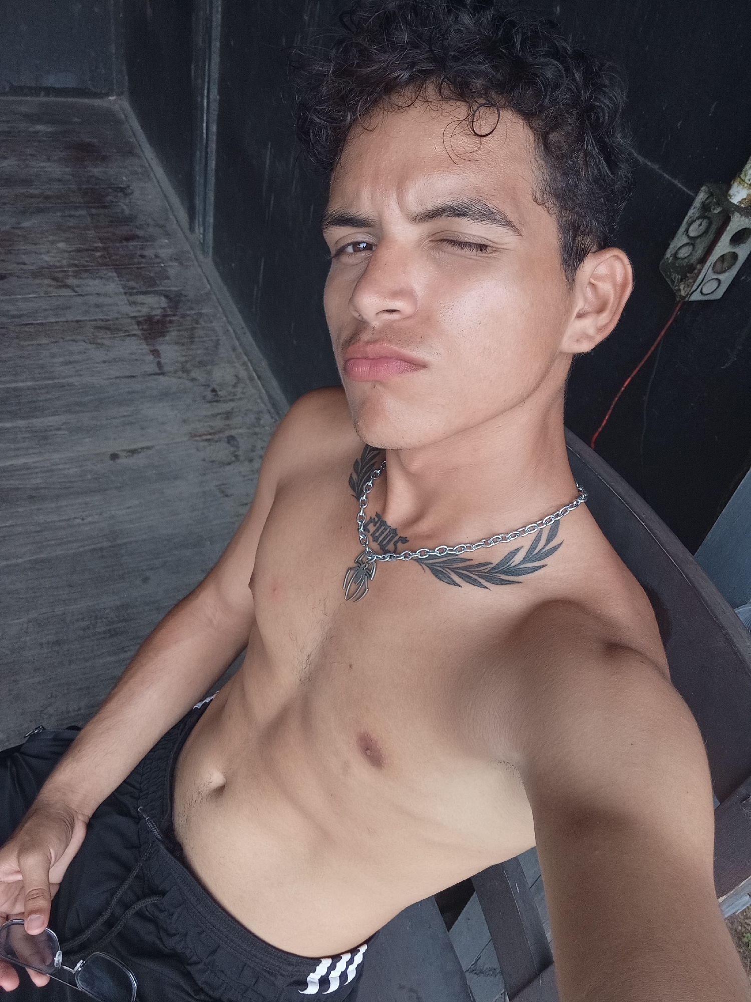 A shirtless, light-skinned man is seated, winking, and looking directly at the camera. He has dark, curly hair, a tattoo on his chest that includes leaves and script, and a silver chain necklace with a spider pendant. He is wearing black athletic shorts with white stripes down the side. He is holding a pair of eyeglasses in his left hand.
The background shows a wooden plank floor and a dark-colored wall. An electrical outlet is visible on the wall.
There is no specific indication of Playa El Agua, Venezuela in the photo.