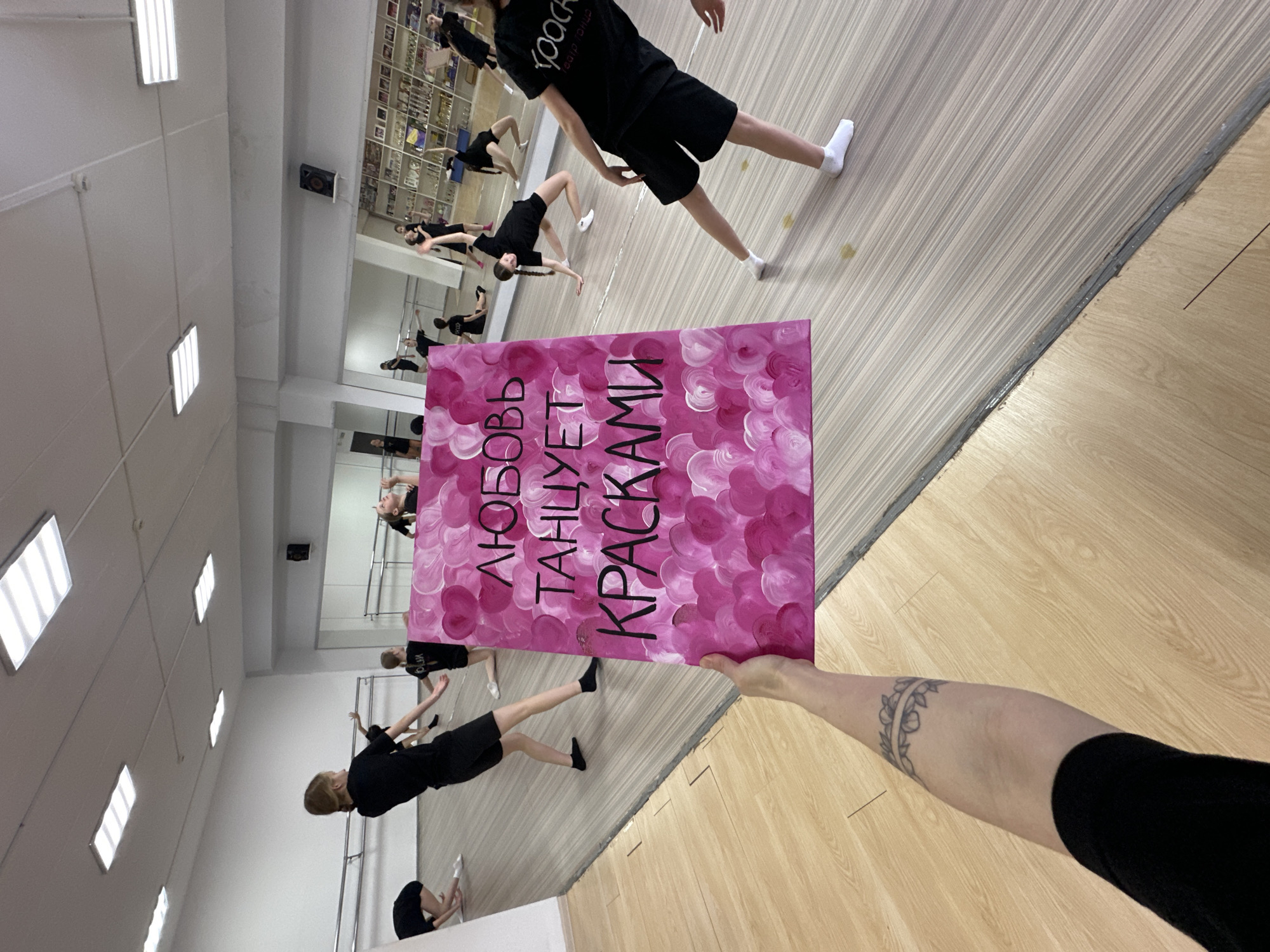 The image is taken inside a dance studio in Votkinsk, Russia. The studio has a wooden floor and white walls, with long, rectangular fluorescent lights on the ceiling. Several dancers, dressed in black, are practicing in the room. A large mirror covers one wall, reflecting the dancers and the studio space. An arm is holding a pink painting with black lettering that reads "ЛЮБОВЬ ТАНЦУЕТ КРАСКАМИ". The arm has a tattoo.