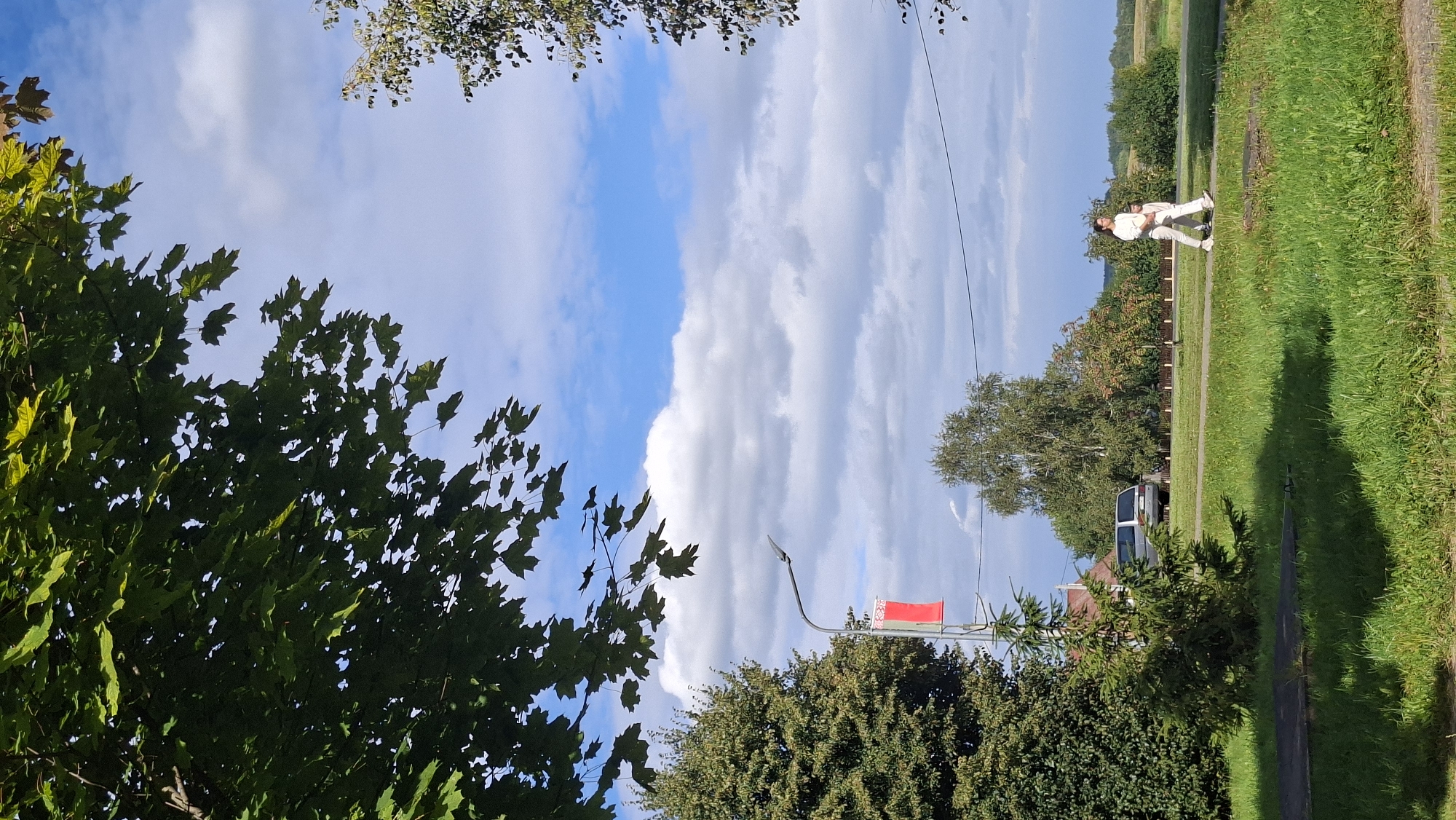 This vertical, outdoor, full-frame shot depicts a sunny day with scattered clouds in the sky over what appears to be a residential area. In the foreground, a patch of green grass is visible, with some shadows cast by unseen objects. A small, conical evergreen tree is positioned to the left of the frame. Further back, a larger evergreen tree stands to the left, and another deciduous tree with light green and some reddish leaves is situated more centrally. A silver van is parked on a road behind a small evergreen tree. To the right of the frame, a person dressed in white, possibly a woman, is walking on a paved path alongside a grassy area. The background features a line of trees and a hint of a rural landscape. A street lamp with a red banner is visible on the left side of the image. The overall scene suggests a peaceful, everyday moment captured in Myadzyel, Belarus.