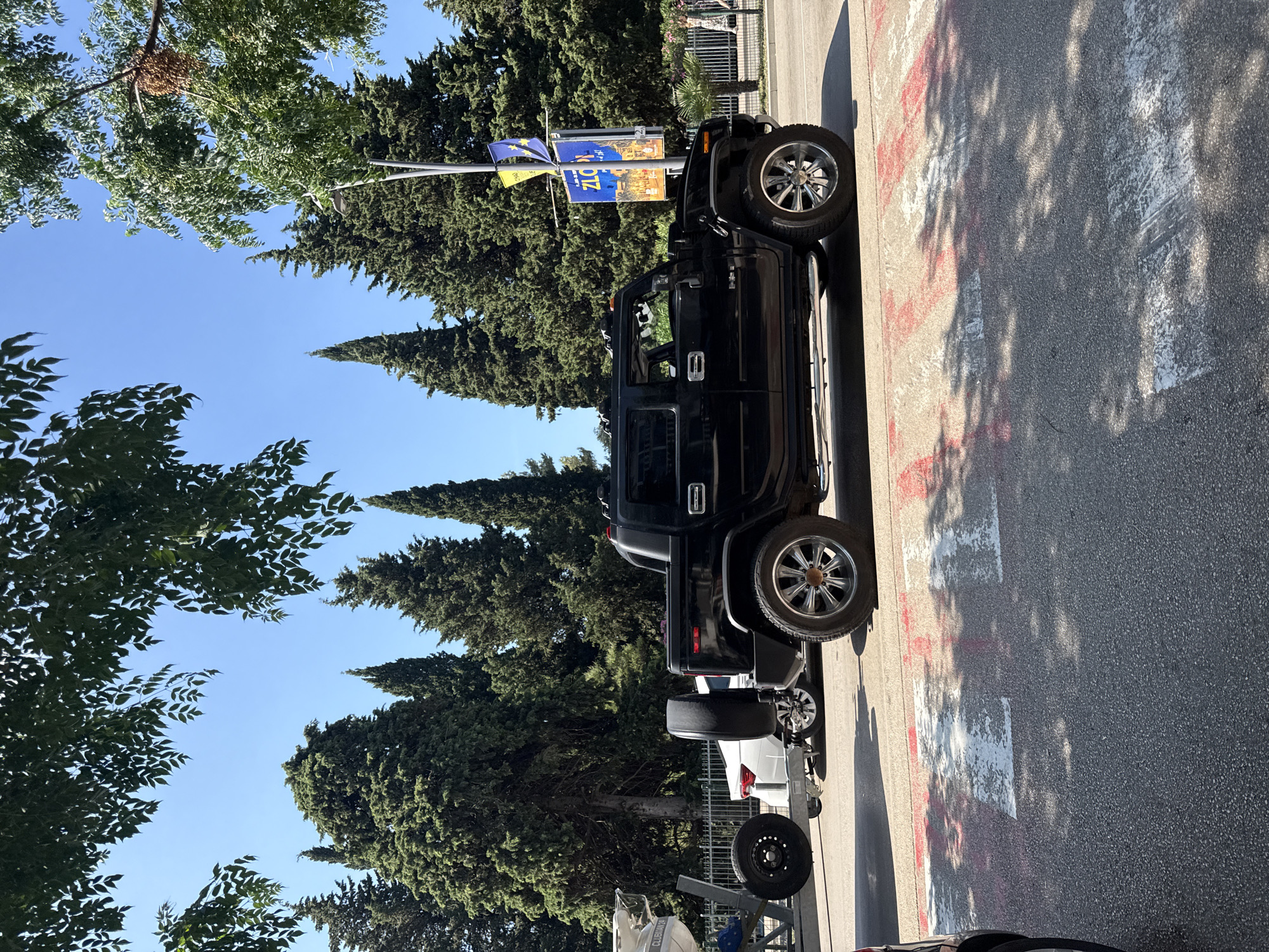 A black Hummer H2 pickup truck is parked on a street in Budva, Montenegro, in front of a line of cypress trees. The truck is viewed from the side, with its front facing left and its rear facing right. The truck has a spare tire mounted on the back. To the left of the Hummer, a white boat on a trailer is visible. The street has a crosswalk marked with white and red paint. In the background, a banner with the text "ZLO" is displayed on a pole. The sky is clear and blue.
