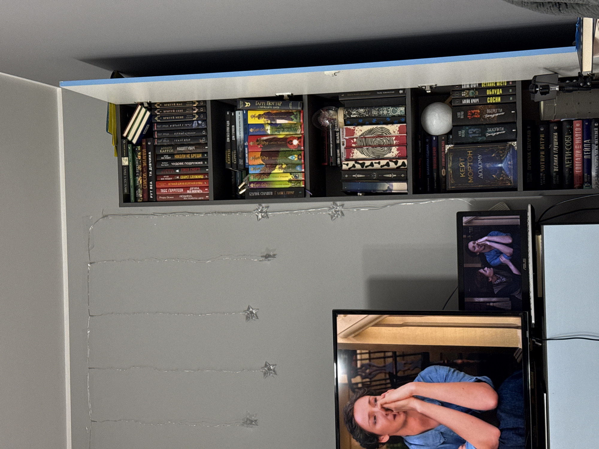 This is a full-frame, eye-level, indoor, medium shot of a room interior. In the foreground, a portion of a television screen displays a scene of a woman with her hands clasped in front of her face. To the right of the television, a laptop displays a similar scene with two women. Behind the electronics, a grey wall is adorned with strings of star-shaped lights. To the right of the wall, an open, light blue cabinet reveals several shelves filled with books. The cabinet's interior is dark grey, and the shelves are packed tightly with books of various sizes and colors. Some books are stacked horizontally on top of others. The overall impression is one of a living room or study area. The image does not provide any specific information or clues about the location within the city of Łódź, Poland.