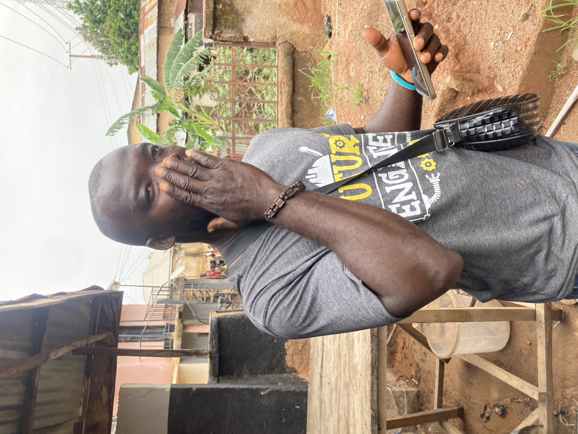 A medium shot shows a man looking at a cell phone in his right hand, covering his mouth with his left hand. He wears a grey t-shirt with black text, and a patterned black bag is slung over his shoulder. He also wears a beaded bracelet on his left wrist. The background shows a partially-constructed wooden platform, a building with corrugated metal roofing, and vegetation. The ground appears to be dirt. The location is Gwong, Nigeria.