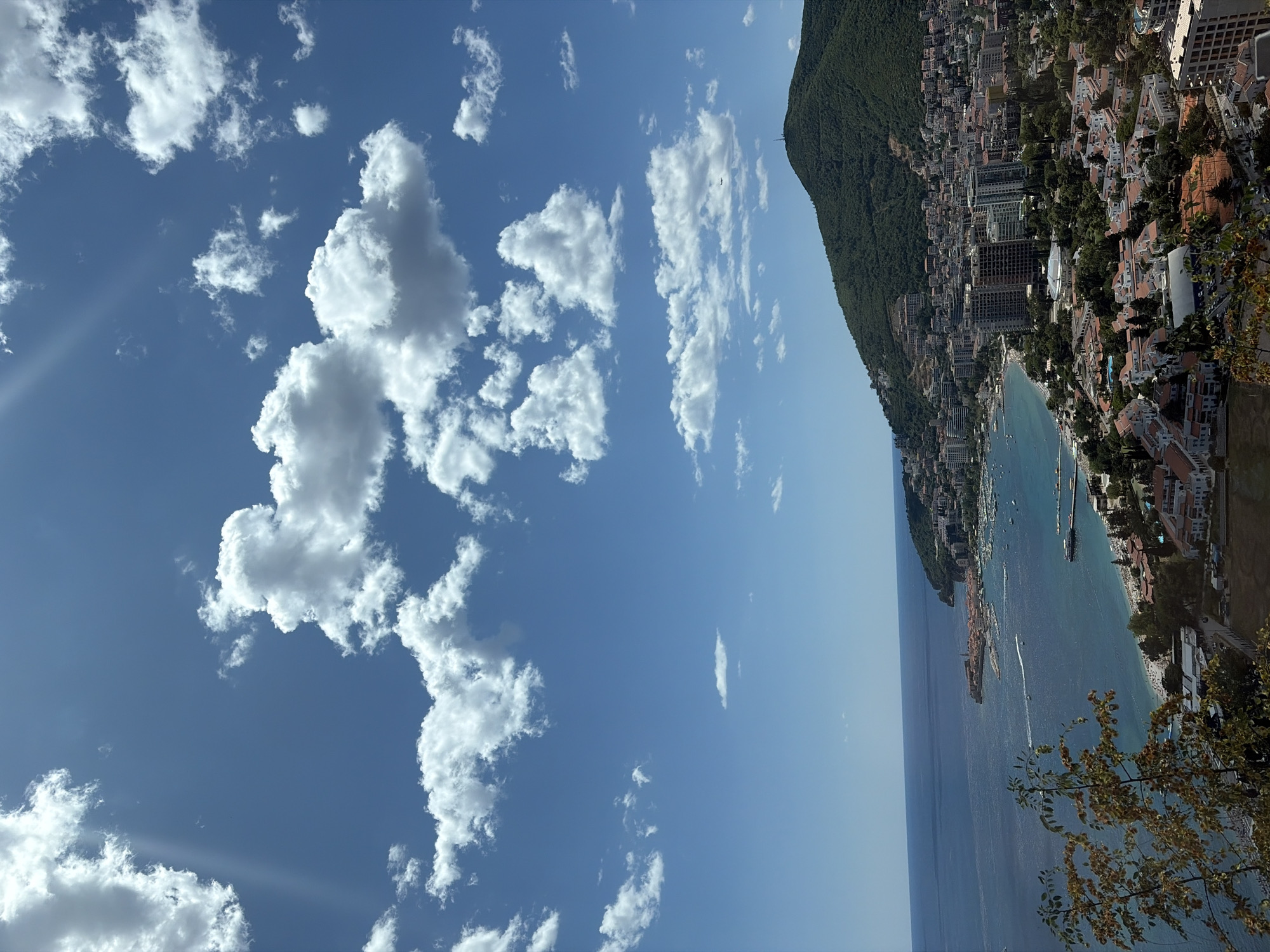 This is a high-angle, full shot of a coastal city on a sunny day, likely taken from a hilltop overlooking the Adriatic Sea. The sky is a bright blue, dotted with fluffy white cumulus clouds, some of which are illuminated by the sun. The sea is a deep blue, with a calm surface reflecting the sky. In the foreground, on the left, is the edge of lush green trees.
The city, identified as Košljun, Montenegro, sprawls along the coastline and up the side of a steep, densely forested green hill that dominates the right side of the image. The architecture is a mix of modern high-rise buildings, some appearing to be under construction, and smaller, reddish-tiled roofs of older buildings. A crescent-shaped bay forms the central feature of the city's waterfront, with numerous boats, including yachts and smaller vessels, moored or sailing in the water. A beach area with umbrellas and people is visible along the shoreline. A prominent breakwater or pier extends into the sea. A small island with buildings, possibly an old town or fortress, is situated within the bay. The forested hill has a communication tower near its summit. The overall impression is one of a vibrant, developed coastal resort area bathed in warm sunlight.