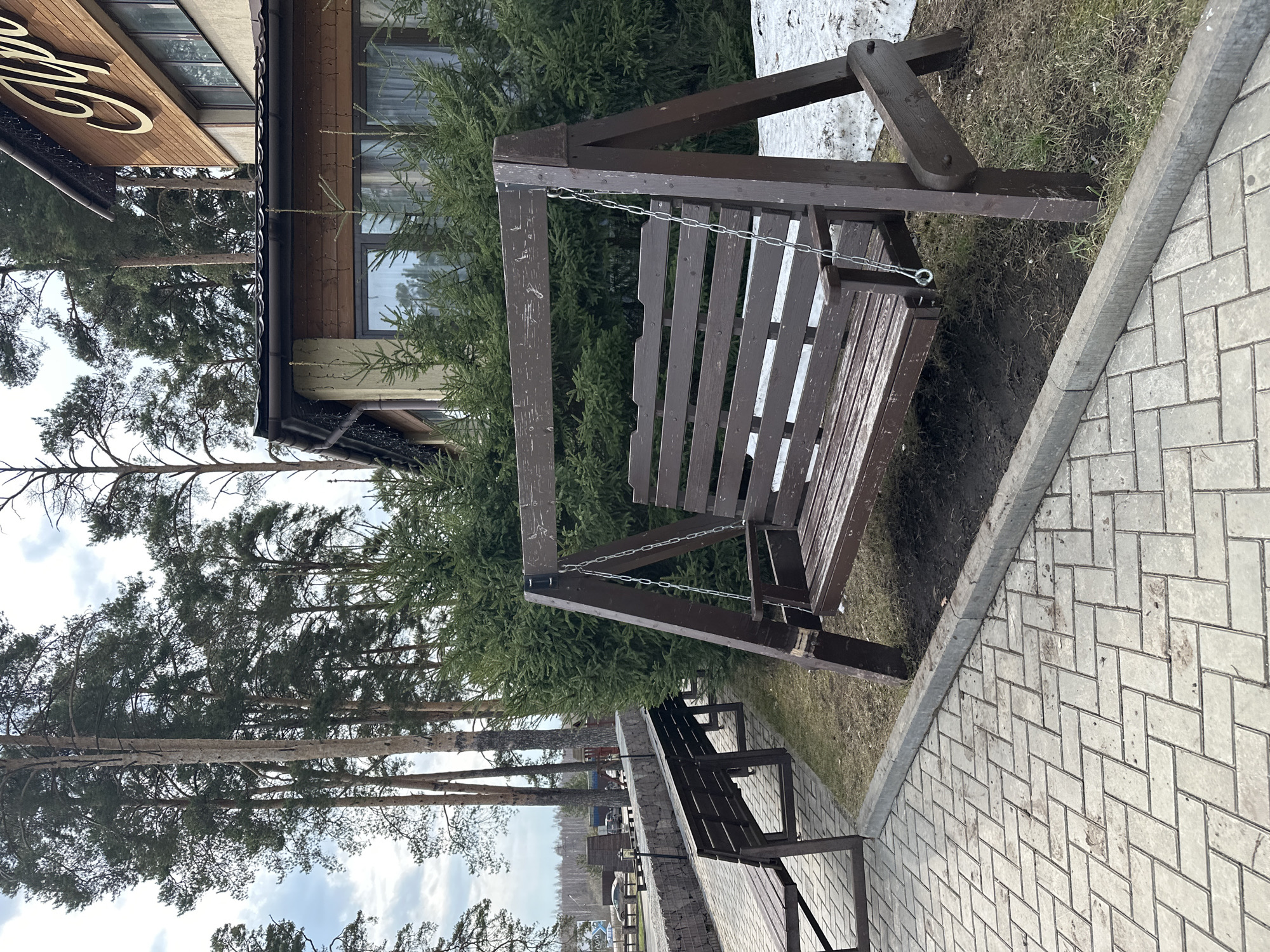 A wooden swing hangs outdoors next to a building and a paved walkway. Behind the swing are evergreen trees, with taller deciduous trees visible in the distance under a cloudy sky. In the lower part of the image, another wooden bench is visible. Based on the location identified as Votkinsk, Russia, it is likely a park or recreational area. There are no people visible.