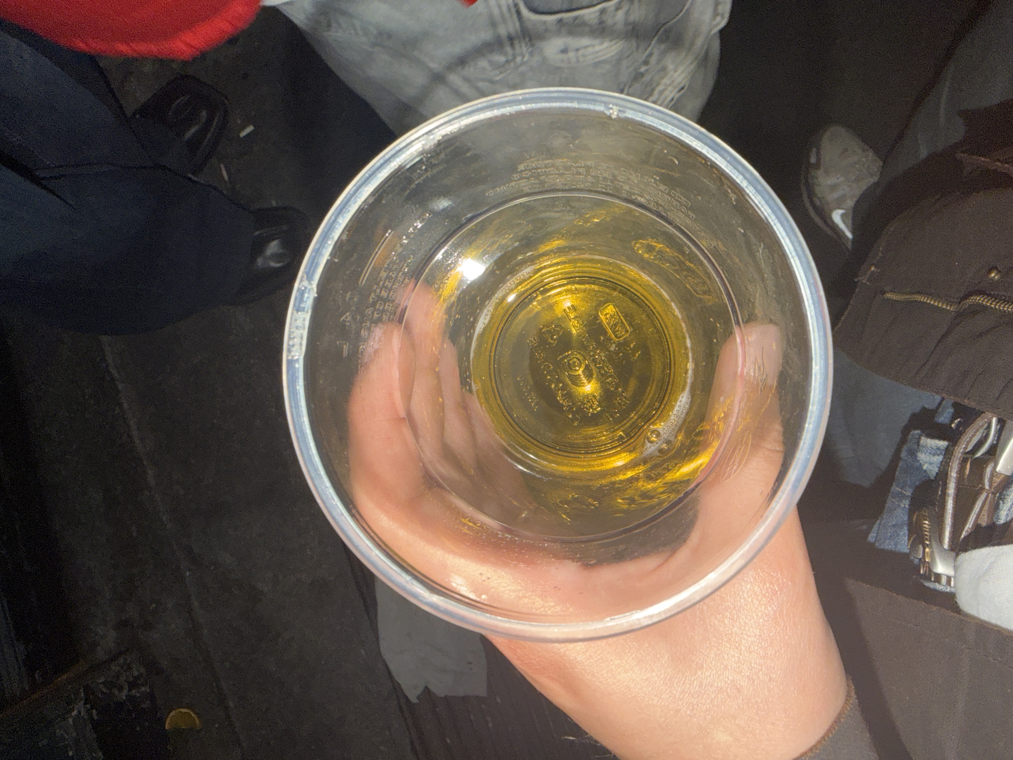 An aerial shot depicts a hand holding a clear plastic cup containing a small amount of yellow liquid. The background includes various articles of clothing: blue jeans, a red garment, black shoes, and what appears to be a dark jacket or coat with a metal zipper. The setting seems to be an outdoor or indoor location with a dark, possibly asphalt surface visible in the bottom left corner. The photo's location as Milan, Italy is not visually evident in the image.