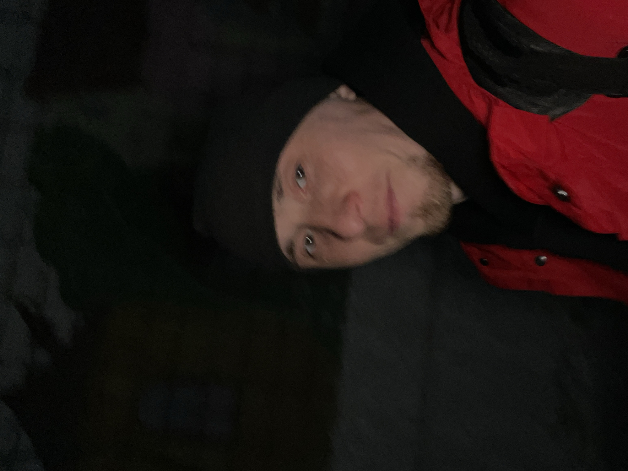 A man is shown from the chest up, turned to his right in a dimly lit setting. He is wearing a black beanie and a red jacket with black accents and silver buttons. He has light skin, light eyes, and a short beard. The environment appears to be outdoors at night, with dark shadows in the background. The location is not discernible as Sarapul, Russia.