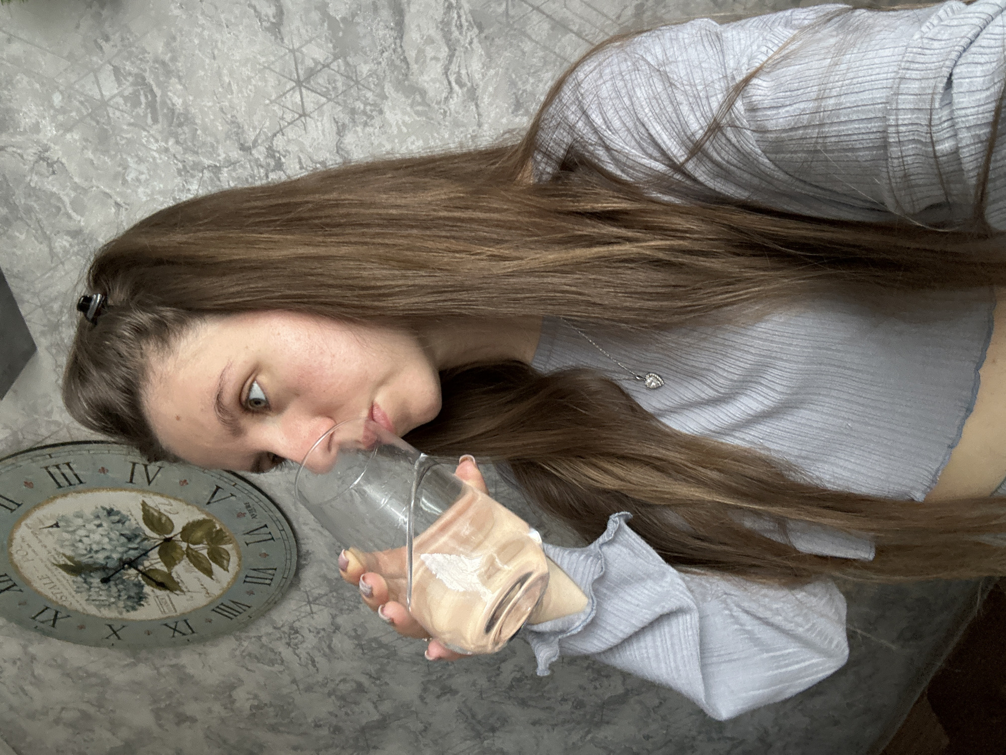 A young woman with long brown hair is drinking from a glass. She is wearing a light blue ribbed top with a ruffled edge. The background includes a gray textured wall and a decorative clock with Roman numerals and a floral design. The location in Klin, Russia, cannot be determined from this image alone.