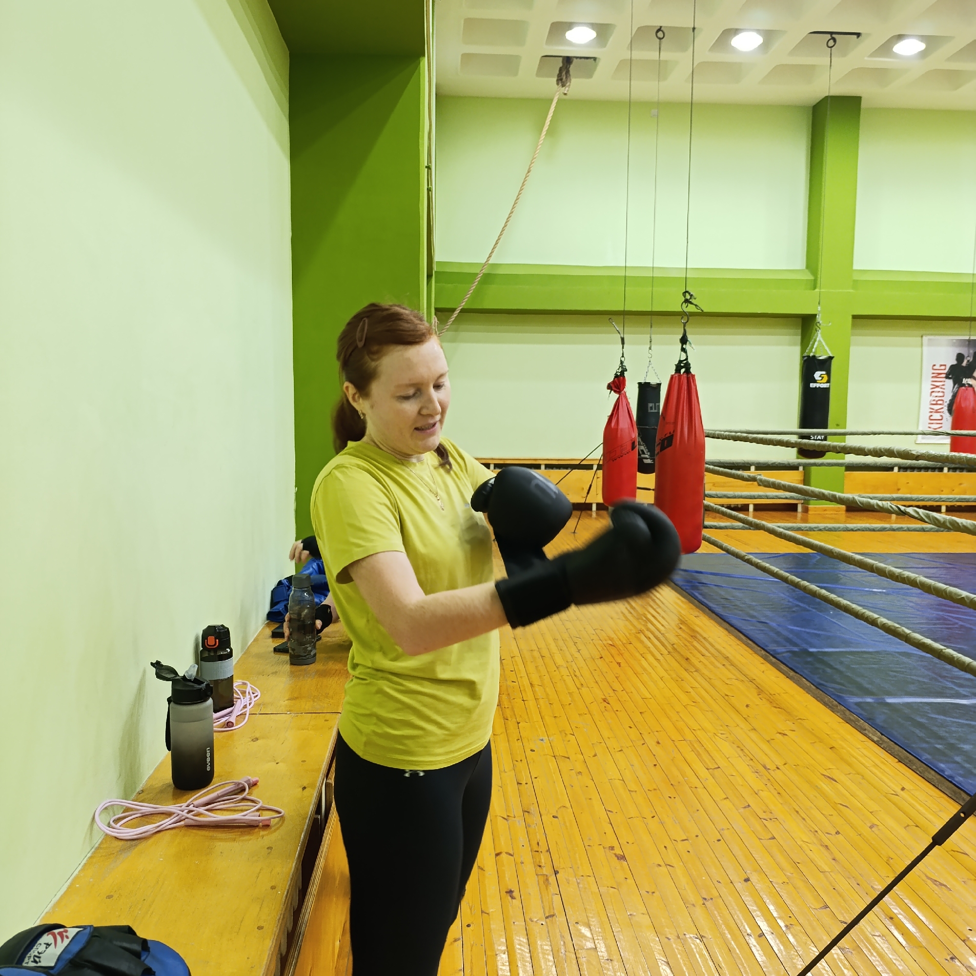 A fair-skinned woman with red hair is putting on black boxing gloves in what appears to be a boxing gym. She is wearing a yellow short-sleeved shirt and black leggings. To her left is a long wooden bench with water bottles, a jump rope, and a blue duffel bag. In the background, there is a boxing ring with red and black punching bags hanging. A "Kickboxing" banner is visible on the wall in the far right corner. The walls of the room are painted a pale green and a brighter green. The floor is wooden. The scene appears to be indoors with artificial lighting. The location is in the city of Unknown, Unknown.