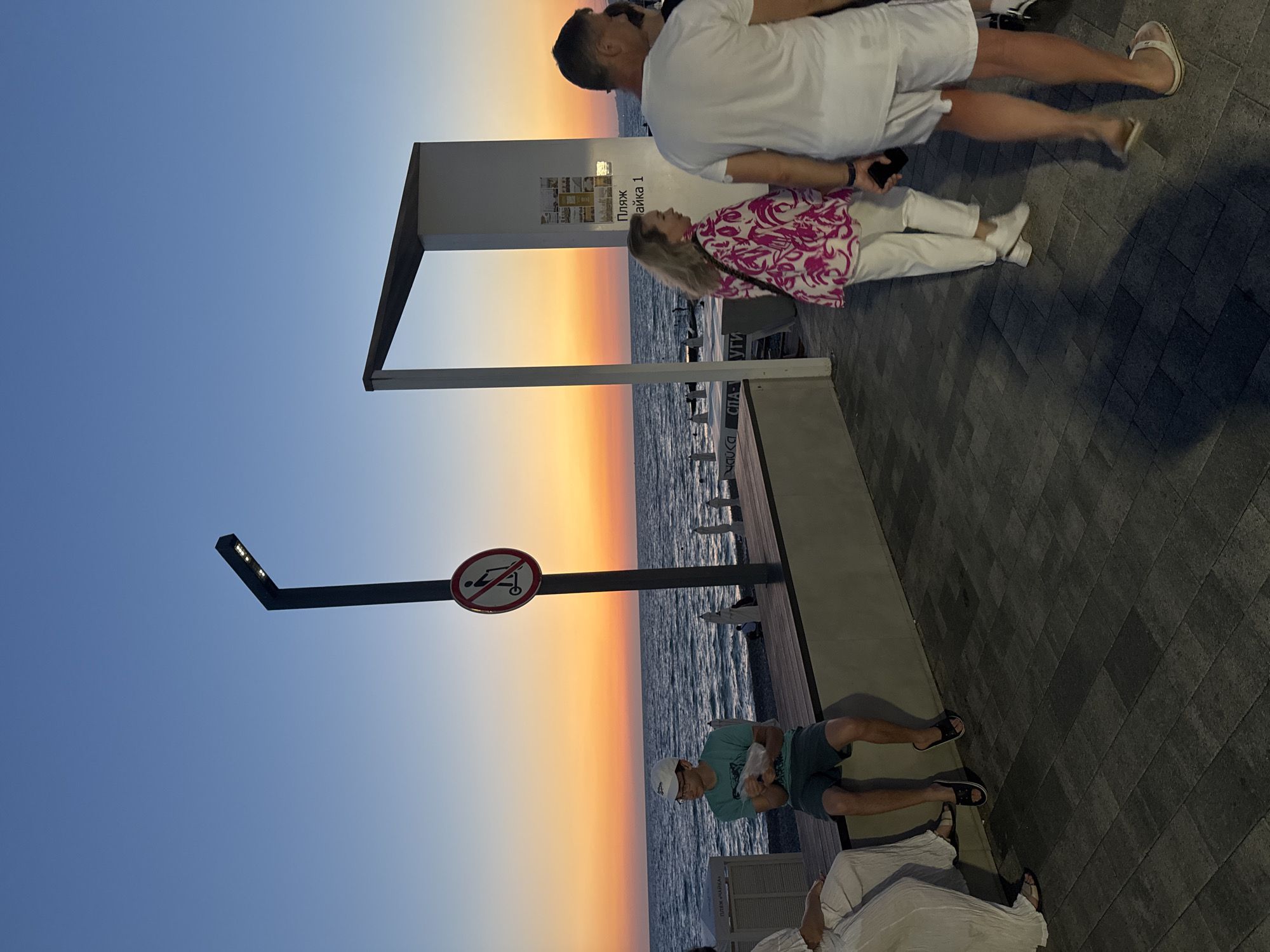 The image captures a dusky scene at the Sochi embankment, likely during the evening. The sky transitions from a deep blue overhead to hues of orange and yellow near the horizon, indicating sunset or sunrise over the Black Sea. The sea itself is visible, with gentle waves rolling towards a pebble beach.
Several people are present, enjoying the ambiance. In the foreground, a young man wearing a white cap, glasses, and a teal t-shirt with dark shorts sits on a wooden bench, holding what appears to be a plastic bag. To his left, a person in a white outfit is partially visible. Further along the embankment, a woman with blonde hair, wearing a pink and white patterned top and white pants, walks with a man in a white t-shirt and white shorts. Both are walking away from the camera.
A modern, minimalist structure with a white pillar and an angled dark roof stands on the right side of the image. A sign with Cyrillic text, "ПЛЯЖ ЧАЙКА 1" (Beach Chaika 1), is affixed to the pillar, identifying the location as Beach Chaika 1. Nearby, a street lamp with multiple LED lights is mounted on a dark pole. A circular red and white sign with a pictogram indicating "no scooters" is attached to the same pole. The ground is paved with rectangular grey tiles. Beach chairs are visible further down the embankment, near the sea.