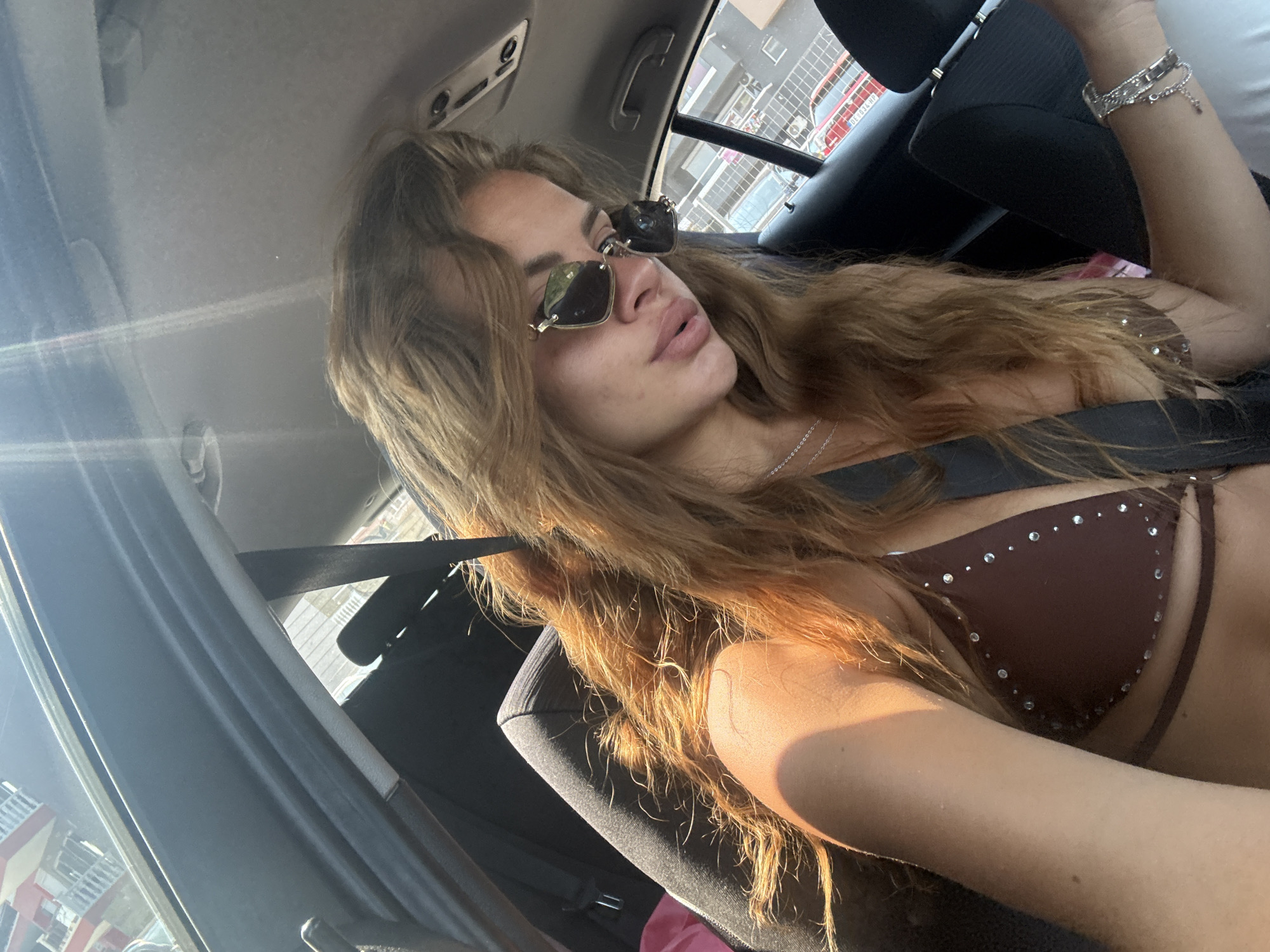 A woman is pictured inside a vehicle, appearing to be in the backseat. She is wearing a brown bikini top adorned with rhinestones and sunglasses. Her hair is long and light brown, falling around her shoulders. The interior of the car is visible, including a seatbelt and part of the car's roof lining. Sunlight streams into the vehicle, creating bright highlights and lens flare. Through the car window, a building with red and white elements is visible, along with other vehicles and urban scenery. The environment suggests a daytime setting, likely in a city, consistent with the provided location of Katerini Municipality, Greece.