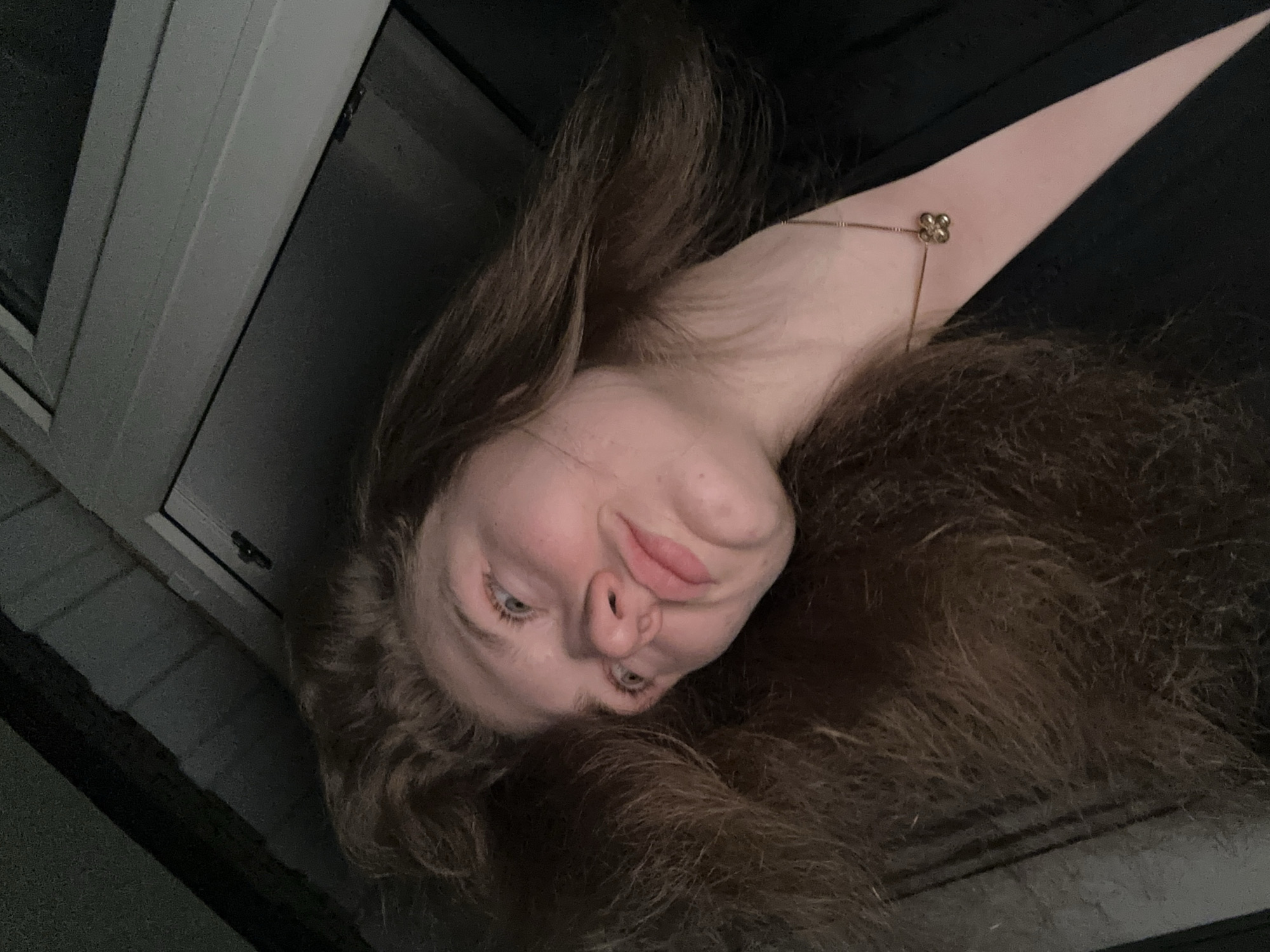 A photograph, taken indoors, presents a woman lying at an angle. Her face is angled towards the viewer with brown hair spread around her. She is wearing a gold necklace. The background appears to be a dark wall, and a window frame is visible to the left. No evidence in the image indicates the location as Kazan, Russia.