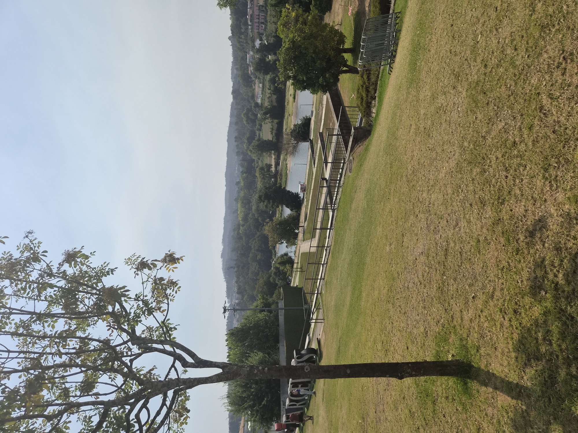 The image shows a view of the Cávado River in Barcelos, Portugal. In the foreground, a tree with sparse leaves stands on a grassy slope. In the middle ground, a large, black, three-dimensional sculpture spells out "BARCELOS." To the right of the sculpture, there is a paved walkway with railings, leading down to the river. The river itself is visible, with a small red and white boat moored on the bank. In the background, a dense forest lines the opposite bank of the river, with some buildings and houses interspersed among the trees. The sky is clear and blue with a few wispy clouds. Two individuals are standing near the sculpture, and several others are walking along the paved path. In the far right of the image, a stack of metal chairs is visible.