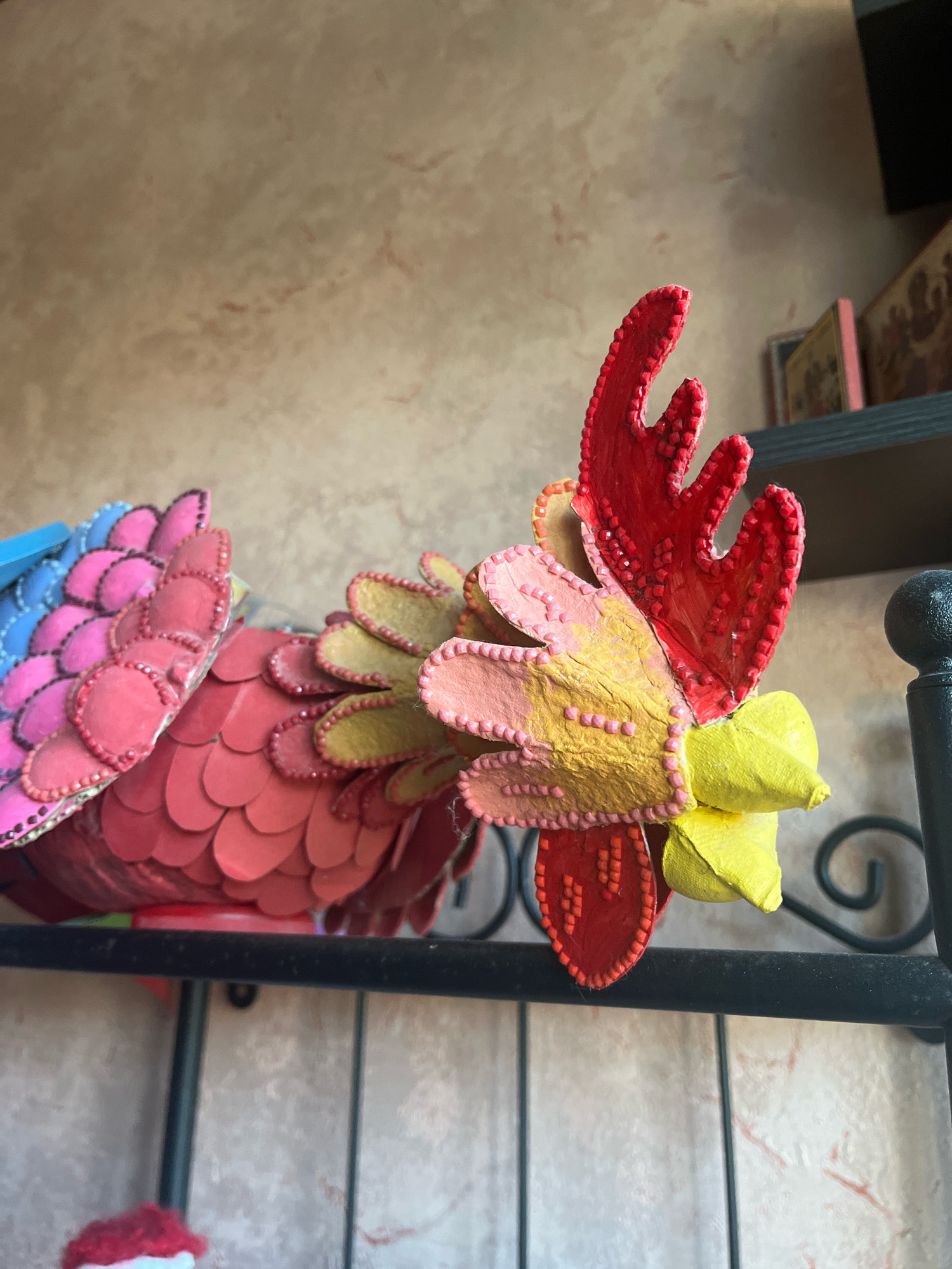 A brightly colored, handcrafted rooster decoration rests on a black metal shelf. The rooster is made of what appears to be paper or felt, with individual scales layered to form its body and tail. The head of the rooster is adorned with red, yellow, and pink accents, resembling a comb and wattles, outlined with small, raised dots. Behind the rooster, a textured wall with subtle pinkish-brown marbling is visible, along with a dark shelf that holds a few framed religious icons. To the right of the rooster, a black metal railing with a scroll design is present. The lighting suggests an indoor environment, possibly a home or a decorative display area. There are no people or animals present in the scene. The location is identified as Vladimirovka, Russia.