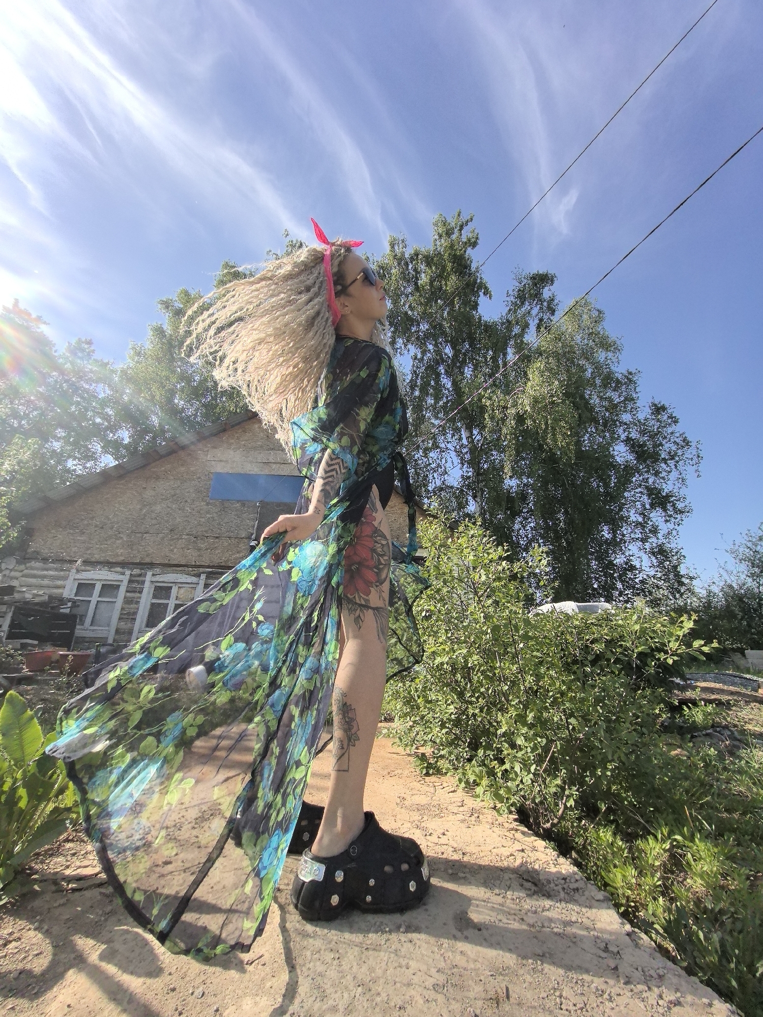 A woman stands outdoors in Noviy, Russia, on a concrete ledge next to lush greenery. She wears a long, semi-transparent floral dress over a black bathing suit and studded black shoes. Her blonde braided hair is styled with a pink headband, and she is wearing sunglasses. The woman is facing away from the camera, her hair and dress are blowing slightly in the wind. Tattoos are visible on her legs and arms. Behind her is a weathered wooden house with white-framed windows, partially obscured by trees. Power lines stretch across the blue sky with wispy clouds. The sun is shining brightly.