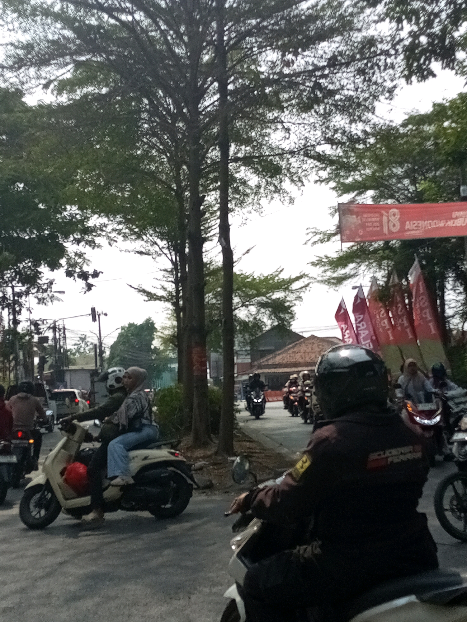 The image shows a street scene in Karangsatria, Indonesia. The foreground is dominated by a person wearing a black helmet and a dark jacket, riding a scooter. Behind them, another scooter is visible with two people on it, one wearing a light-colored hijab and a patterned shirt, and the other wearing a dark jacket and blue jeans. In the background, several other scooters and motorcycles are on the road, some with riders wearing helmets. Trees with dense foliage line the side of the road, and a red banner with white text and the number "18" is visible hanging across the street. Several red flags with white lettering are also displayed. The sky is overcast. The overall impression is a busy street with ongoing traffic.