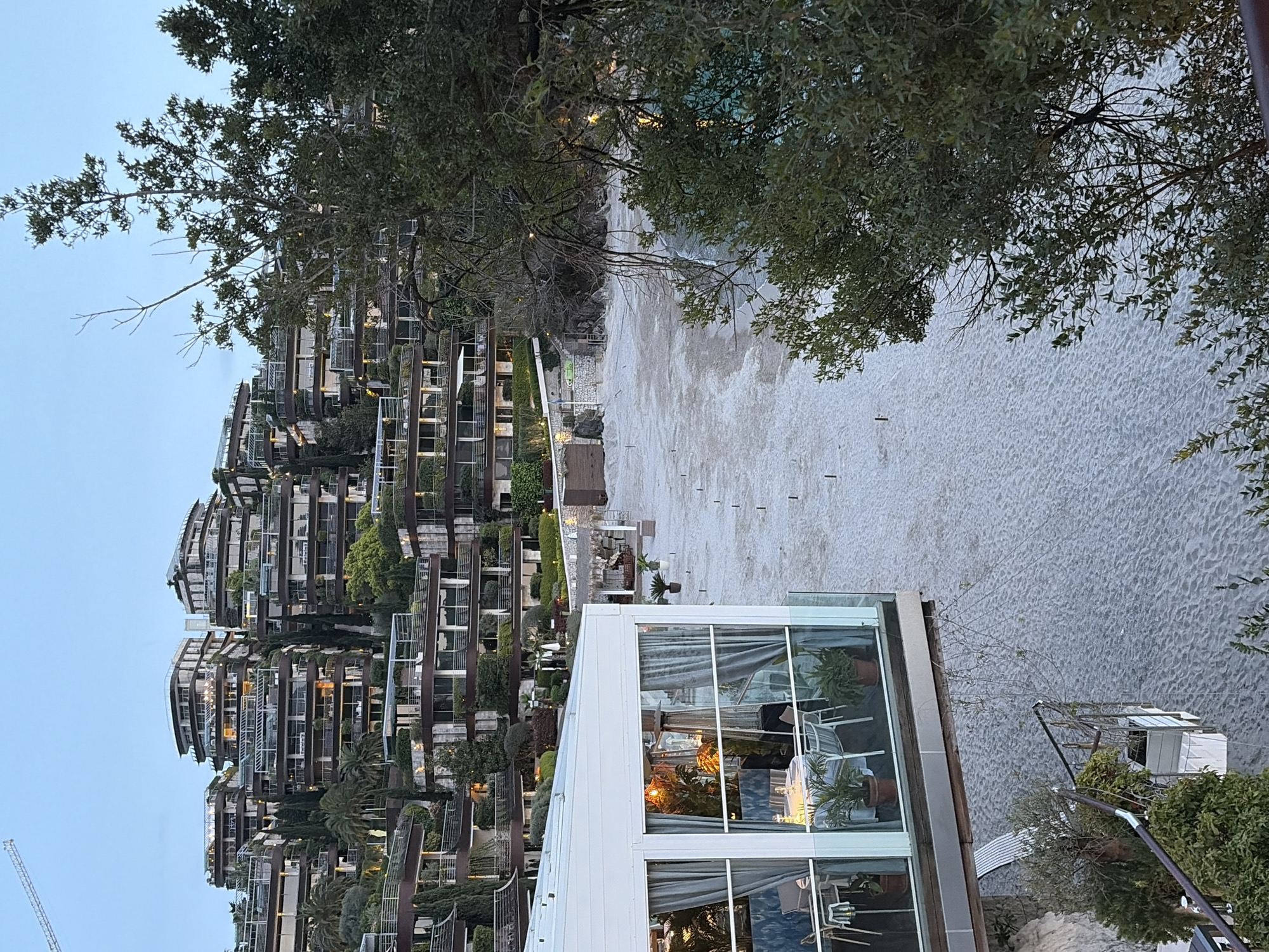 The image depicts a view of a high-end building complex cascading down towards a beach in Budva, Montenegro. The complex features multiple floors with balconies and greenery. A modern, glass-walled structure sits in the foreground, reflecting interior furnishings. The beach appears empty save for regularly spaced markers. Above, there are green trees. A construction crane can be seen on the left side of the building complex. The sky is a pale blue, suggesting dusk or dawn.