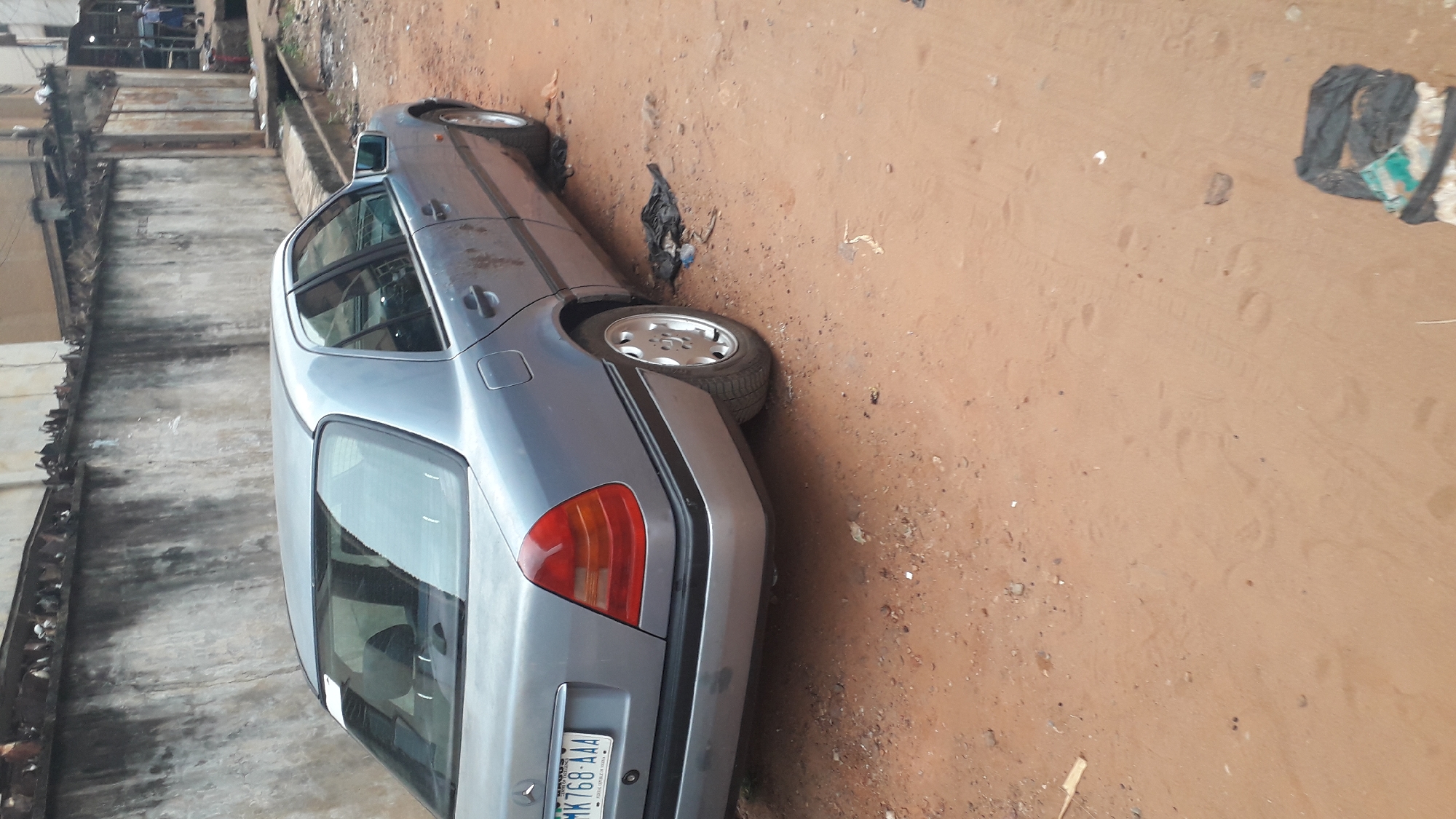 The image shows a gray sedan parked on a dirt shoulder in Obosi, Nigeria. The back of the car is closest to the camera, displaying its license plate. The environment is littered with debris and trash. To the left, there is a concrete wall of a building and the surrounding area appears to be a residential or commercial street. There are no people visible in the picture. The ground is dirt and soil.