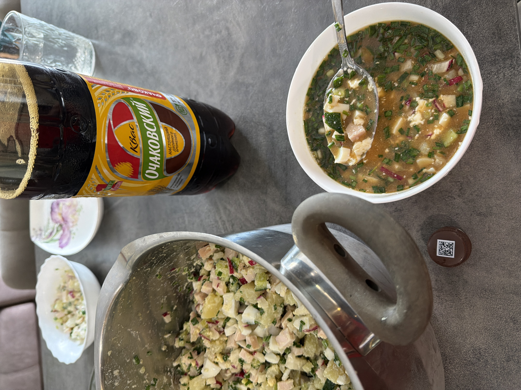 The image shows a table setting with food and drink. A bottle of "Ochakovskiy" kvass is prominently displayed. Kvass is a traditional Slavic and Baltic beverage made from rye bread. There is a bowl of okroshka, a cold soup popular in Russia and Ukraine, made with chopped vegetables, meat, and kvass. A large pot containing the chopped ingredients for okroshka is also visible. Additionally, there's a glass, a small bowl of okroshka ingredients, and a small plate. The image does not contain any evidence to suggest that it was taken in Birobidzhan, Russia. There are no people present in the image.