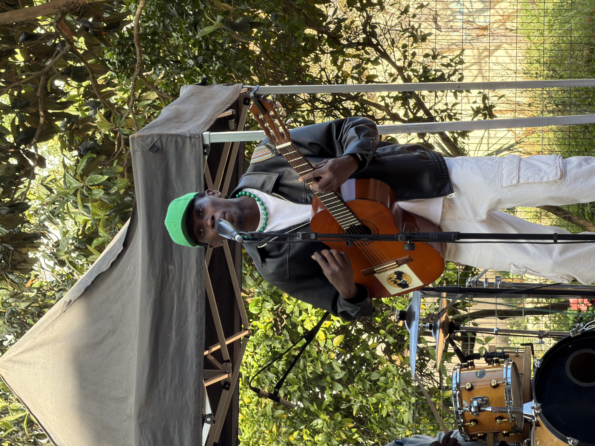 A man is playing an acoustic guitar on a stage outdoors, likely in Lisbon, Portugal, based on context. He is wearing a green cap, a black leather jacket with colorful trim on the shoulders, a white t-shirt, a green beaded necklace, and white cargo pants. A microphone stands in front of him. To his right, a drum set is visible. The stage is covered by a grey canopy. The background is filled with trees and greenery, suggesting a park or garden setting. A metal railing is behind the man.