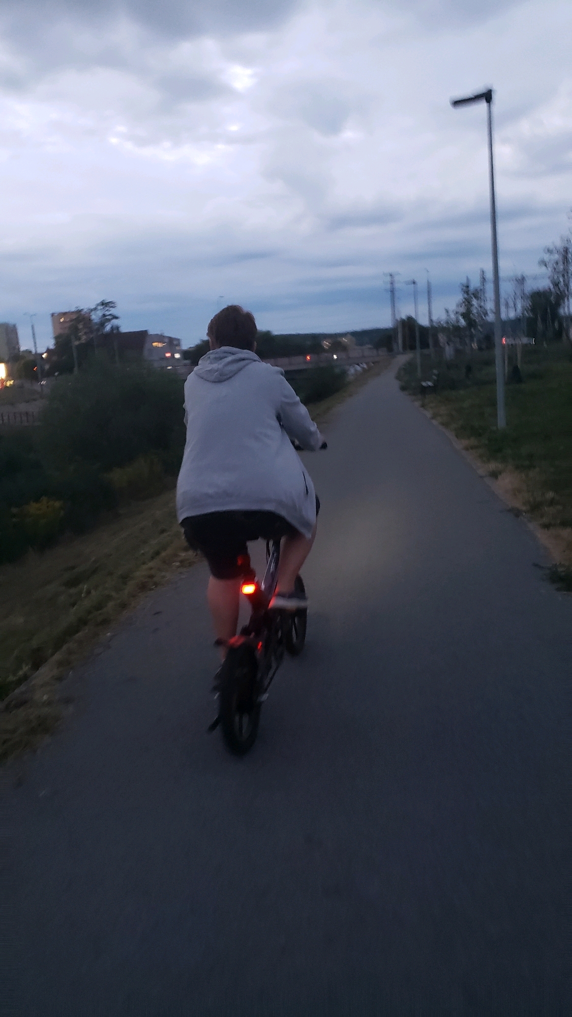 The image depicts a person riding an electric scooter on a paved path alongside a grassy embankment. The person is viewed from behind, wearing a light-colored hooded jacket and dark shorts. The scooter has a red rear light illuminated. In the background, a street lamp stands on the right side of the path, and further in the distance, buildings and trees are visible. The sky is cloudy and overcast, suggesting it is either early morning or late evening. The scene appears to be in an urban or semi-urban park area. While the specific location in Prešov, Slovakia, cannot be definitively identified from the visual elements alone, the presence of urban infrastructure and natural elements is consistent with parkland or a recreational path within a city.