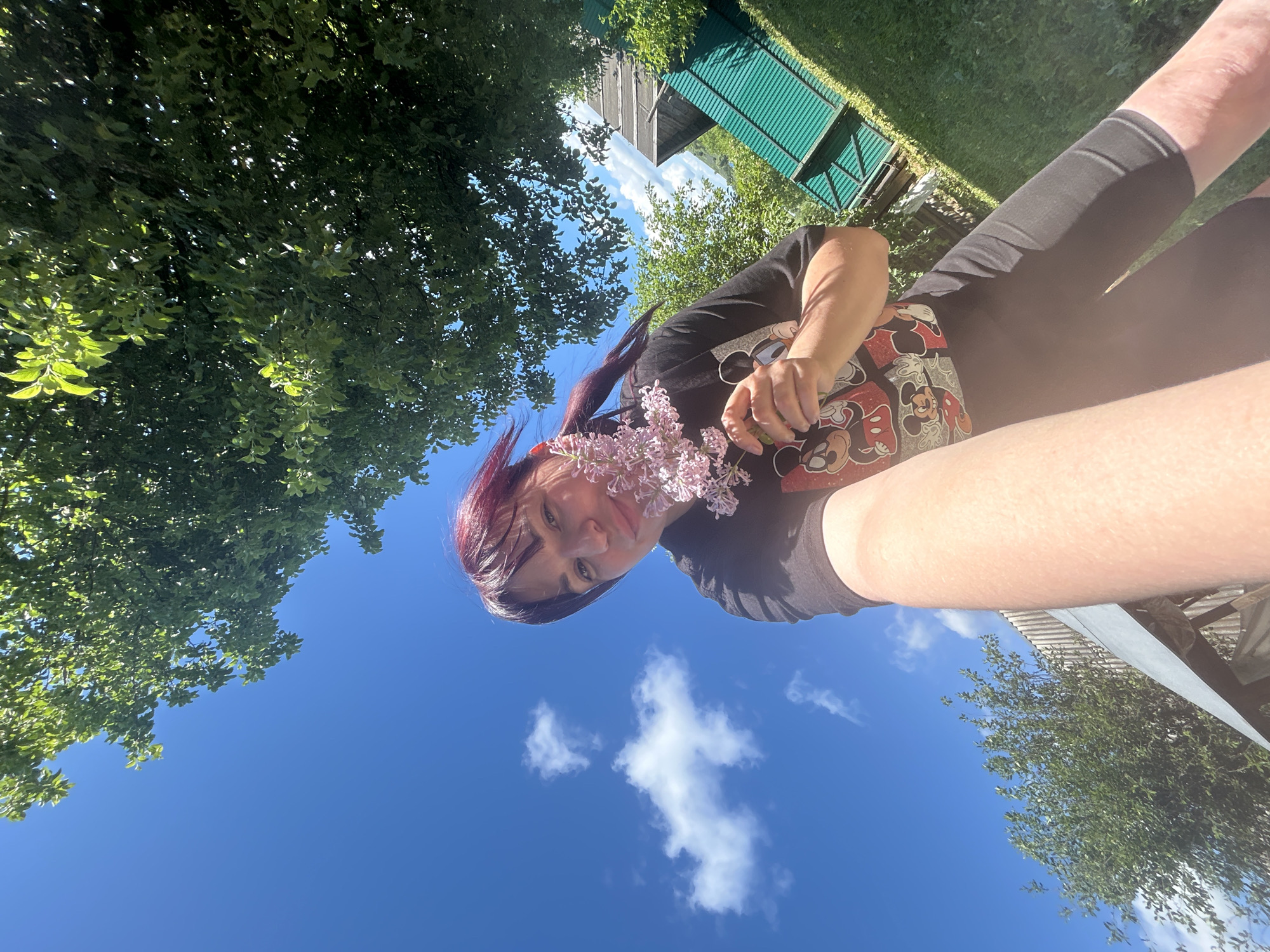 A young woman with dark hair and purple highlights takes a selfie outdoors, holding a sprig of light pink flowers to her face. She is wearing a black t-shirt with a Mickey Mouse graphic and black shorts. The background features a bright blue sky with scattered white clouds, green trees, and some buildings with green corrugated metal roofs. The scene appears to be in a rural or suburban area, possibly within the city of Fokino, Russia. The perspective is low-angle, looking up towards the sky.