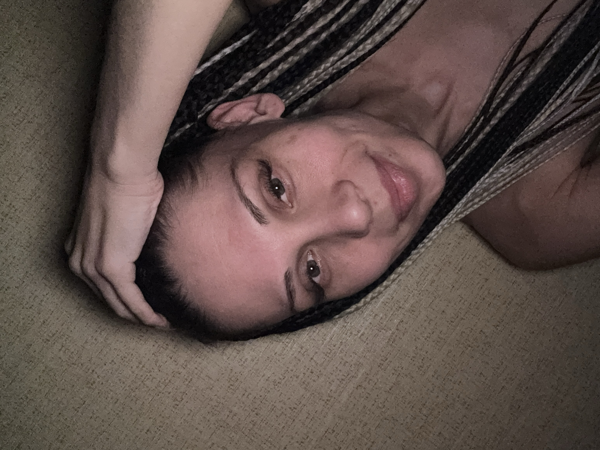A woman with dark and light-colored braids is shown in a close-up shot, lying on a textured surface, possibly a sofa or carpet, in an indoor setting in Rusanovo, Russia. Her head is turned towards the viewer, with her left arm bent and hand resting near her head. She has a slight, relaxed smile on her face, and her brown eyes are looking directly into the camera. Her complexion appears clear, and she has visible eye makeup, including eyeliner and mascara, along with full lips. The lighting in the image is dim, suggesting it might be evening or an interior space with subdued illumination. The overall mood conveyed is relaxed and calm. No visible text is present in the image.