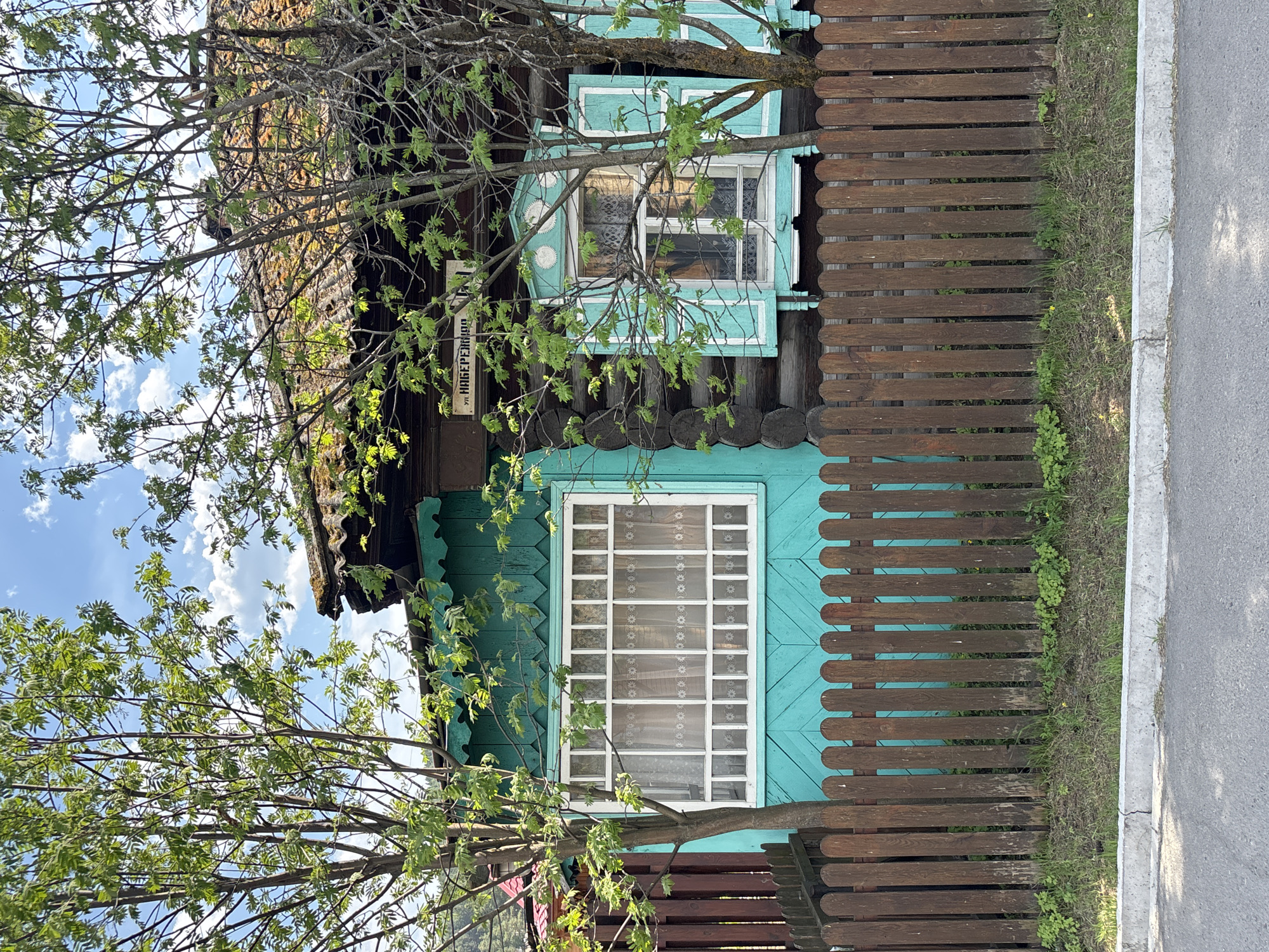 The image depicts a wooden house in Овсянка, Russia. The house is painted in a blue-green color and has white framed windows with lace curtains. A dark brown wooden fence runs along the side of the house, bordering a street. A tree with sparse green leaves partially obscures the view of the house. A sign that reads "УЛ. НАБЕРЕЖНАЯ" is also visible on the house. The sky is visible through the tree branches, with scattered clouds. The street is made of asphalt.