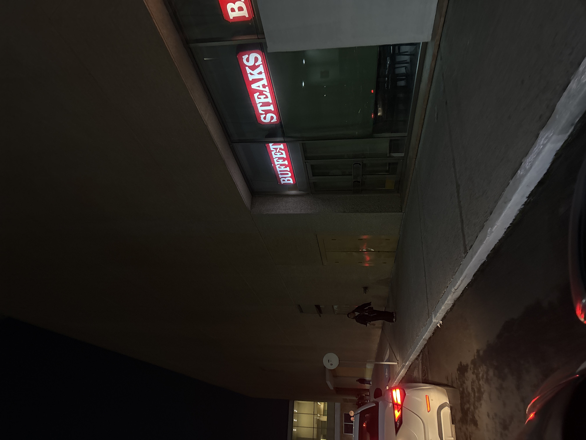 At night in Metepec, Mexico, a restaurant storefront labeled "Buffet Steaks B" faces a sidewalk. A person walks along the sidewalk. A white car is parked along the curb, its tail lights illuminated. The building's facade is predominantly neutral in color, contrasting with the black of the night sky.
