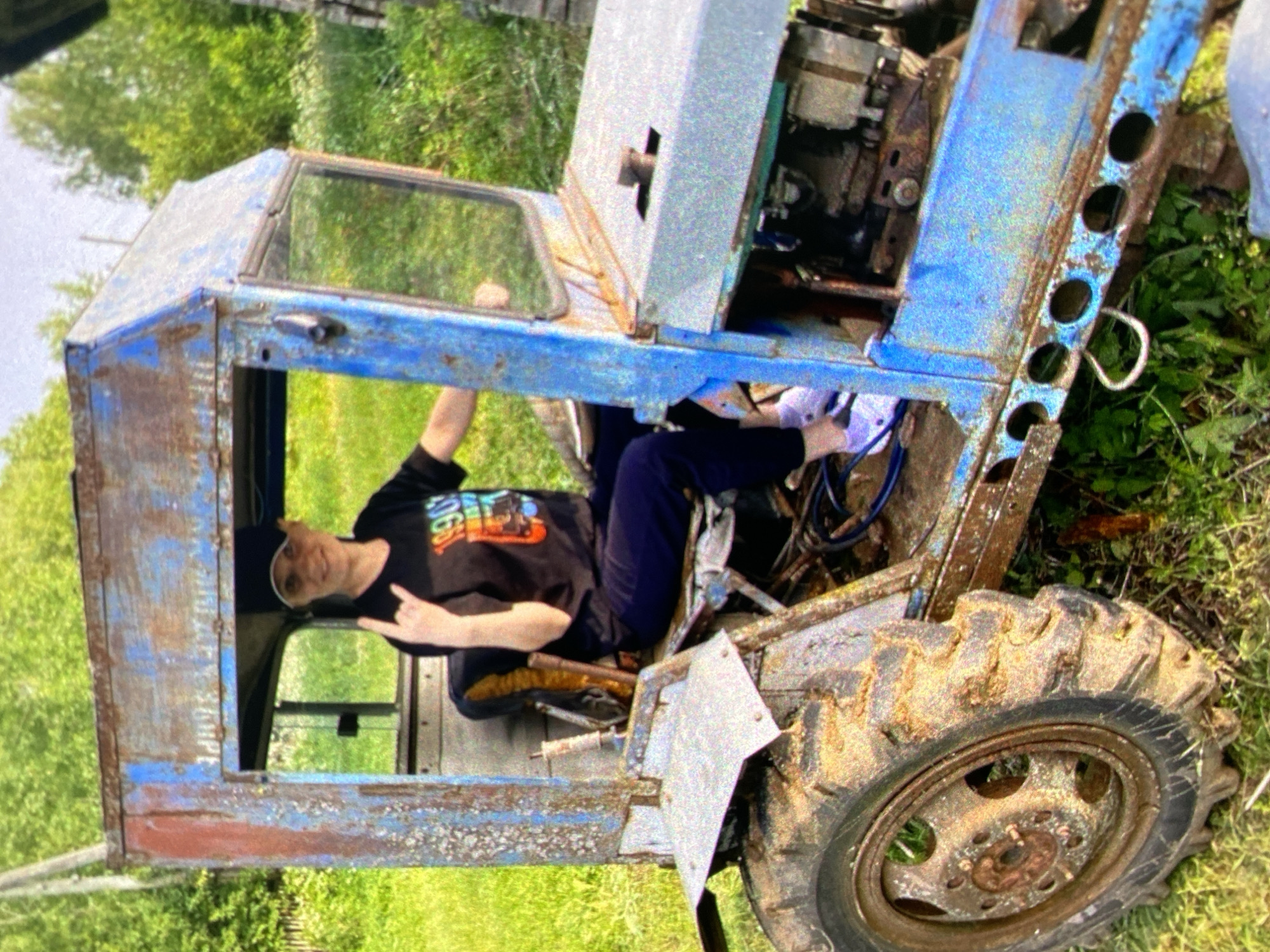 A person is seated inside a rusty blue tractor cab, which appears to be outdoors in a grassy area. The person, wearing a black t-shirt and dark pants, is gesturing with their hand. The tractor appears old and worn, with visible rust. The background includes green trees. This image doesn't provide location information. It is not possible to identify if this photo was taken in Saint Petersburg, Russia.